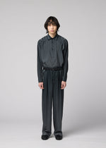 A person with medium-length dark hair stands against a plain gray background, wearing the DAWN RAY dark, loose-fitting button-up shirt and matching pleated pants from IM MEN, which feature a subtle gradation pattern, paired with black shoes.