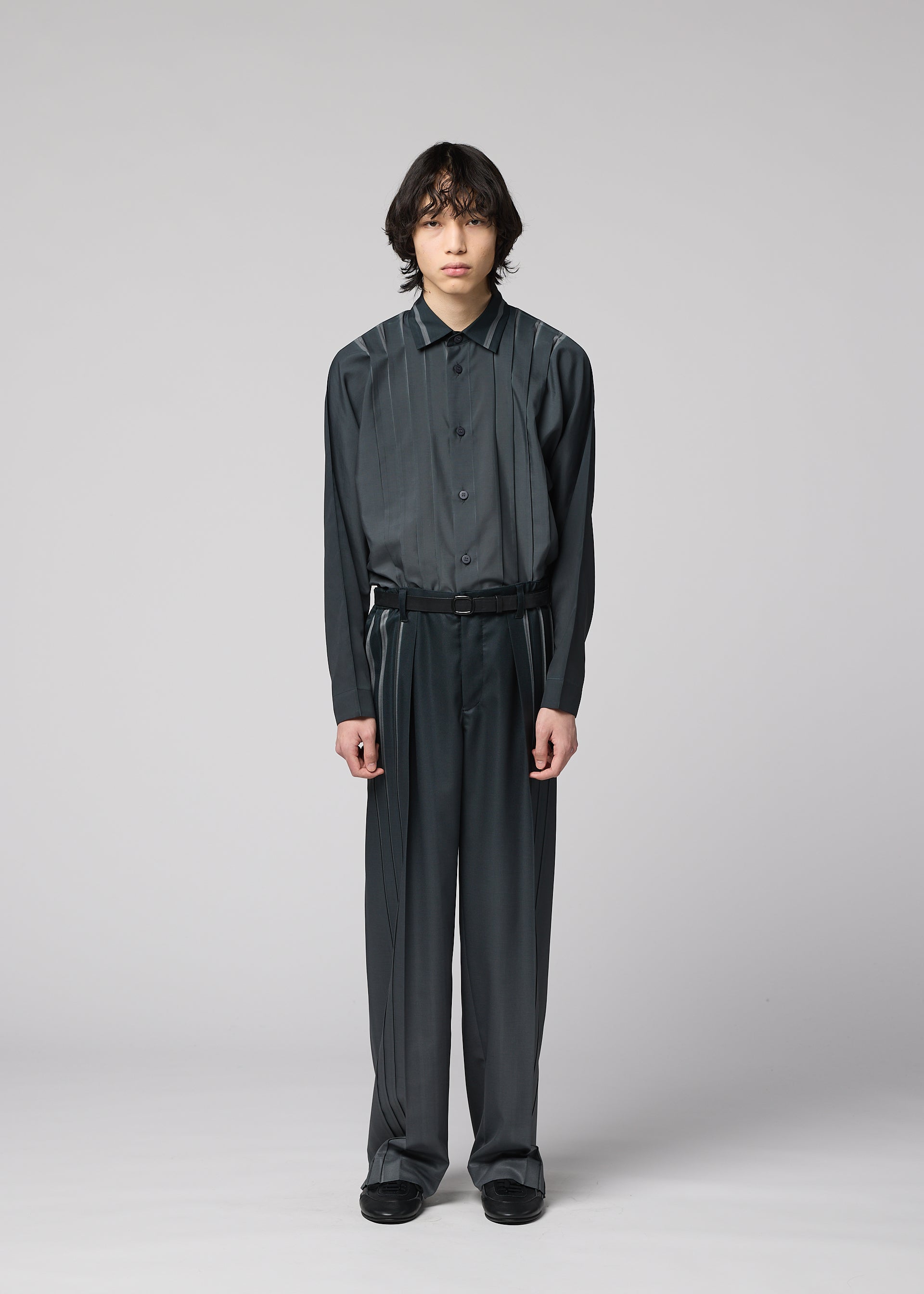 Zoomed view A person with medium-length dark hair stands against a plain gray background, wearing the DAWN RAY dark, loose-fitting button-up shirt and matching pleated pants from IM MEN, which feature a subtle gradation pattern, paired with black shoes.