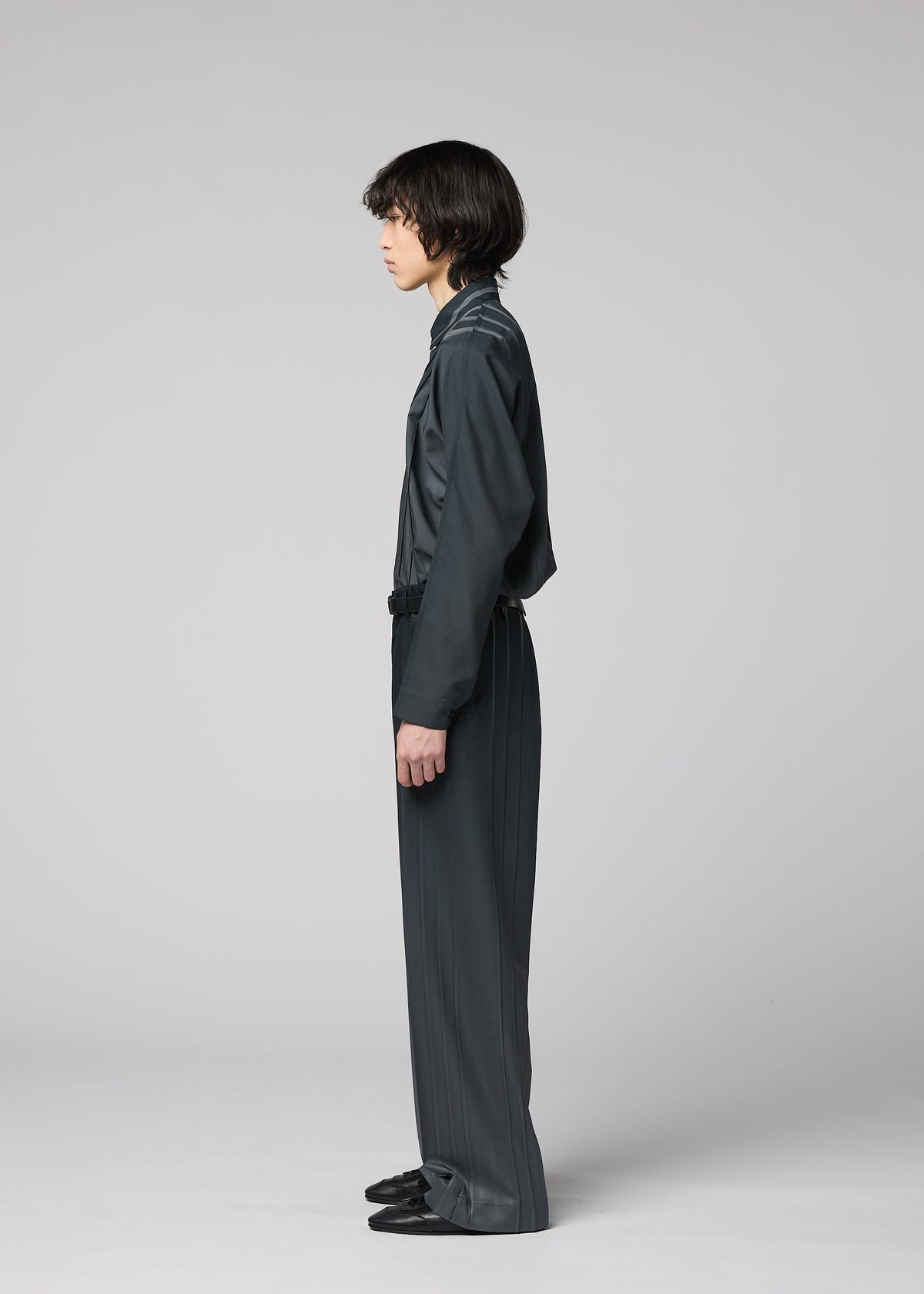 A person with medium-length dark hair stands in profile against a plain light gray background, wearing the DAWN RAY long-sleeve top and wide-leg pleated pants by IM MEN in a loose-fitting, dark gray ensemble. view 5 of 7