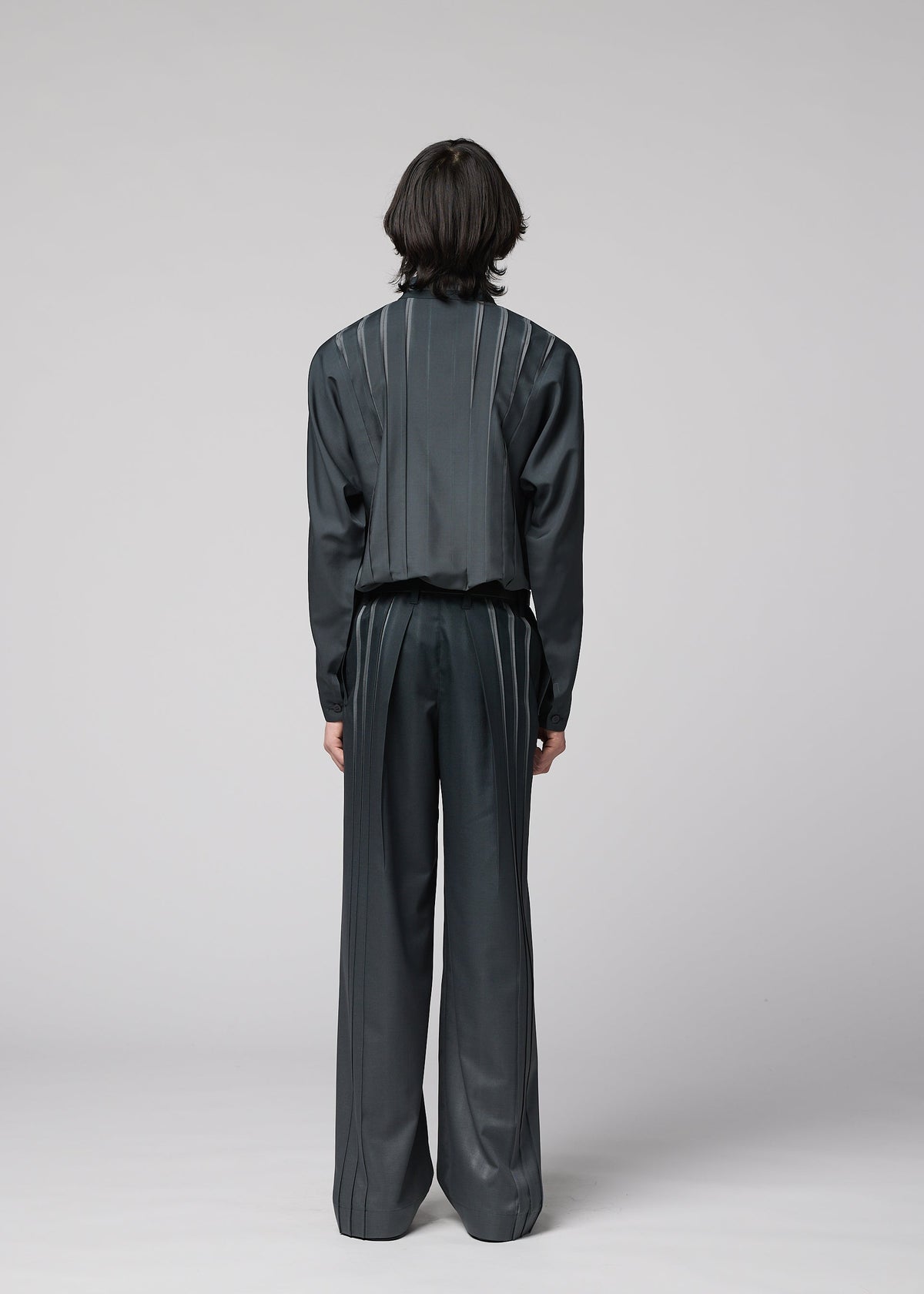 A person with medium-length dark hair stands facing away, wearing the IM MEN DAWN RAY dark, loose-fitting suit featuring vertical pinstripe details and transfer-printed pleated pants, set against a plain light gray background. view 6 of 7