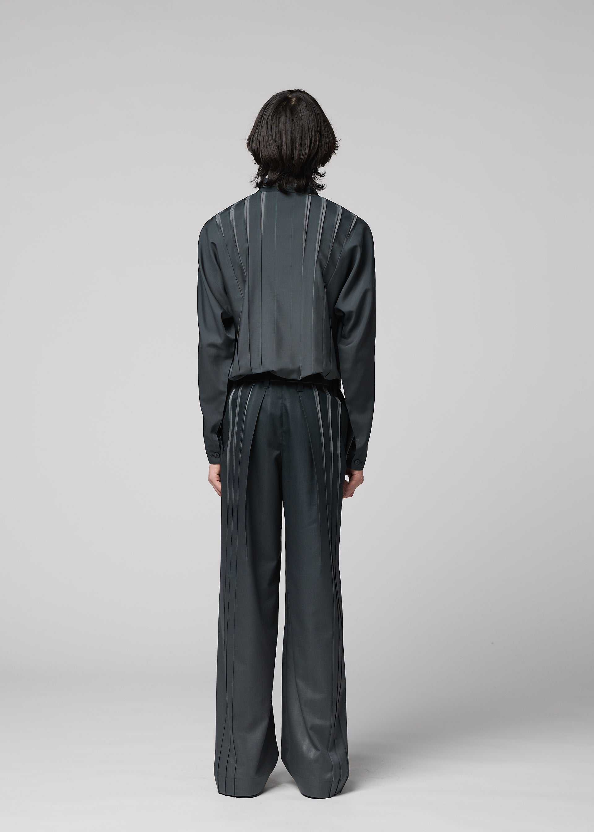 Zoomed view A person with medium-length dark hair stands facing away, wearing the IM MEN DAWN RAY dark, loose-fitting suit featuring vertical pinstripe details and transfer-printed pleated pants, set against a plain light gray background.
