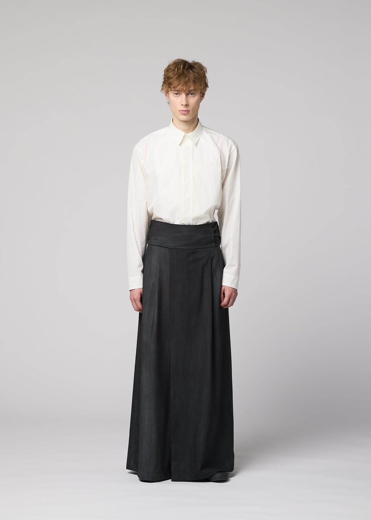 Zoomed view A person stands facing forward in IM MEN’s GRADATION WOOL, featuring a white button-up shirt tucked into long, dark pants. Their short, tousled hair contrasts with the plain light gray background.