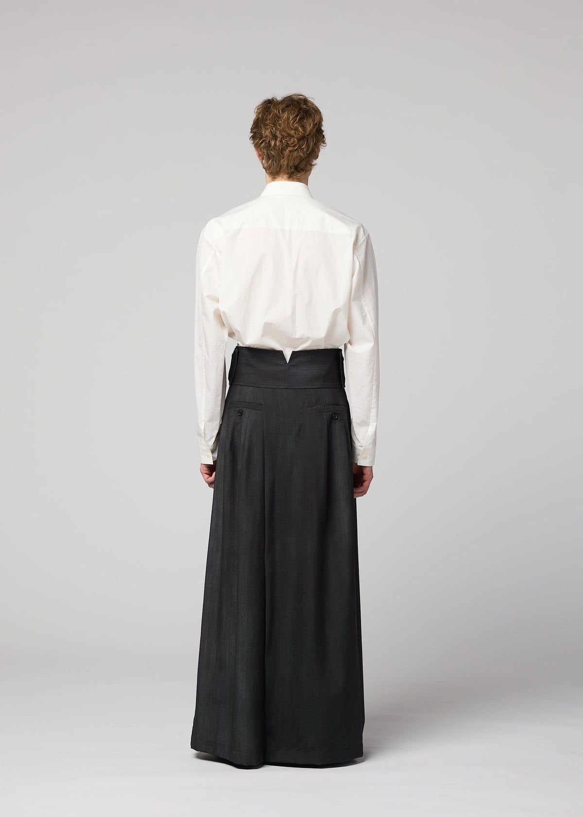 Zoomed view Person standing with back to camera, wearing a white long-sleeve shirt tucked into IM MEN GRADATION WOOL high-waisted, wide-leg pants against a plain light gray background.