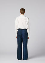 A person with short hair faces away, wearing a white long-sleeve shirt tucked into loose blue GRADATION WOOL pants from IM MEN. The background is plain, light gray.