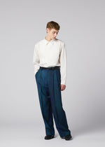 A person stands before a plain background, wearing a white long-sleeve shirt tucked into high-waisted GRADATION WOOL blue plaid pants from IM MEN, styled with a black belt and black shoes.