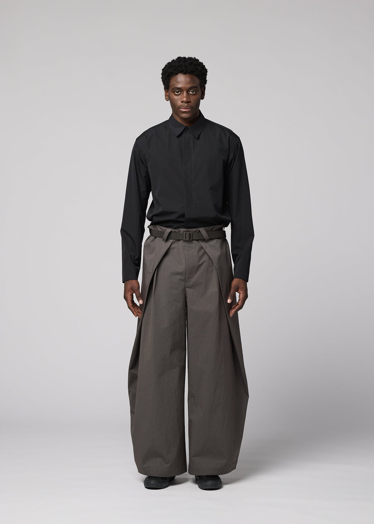A person stands facing forward against a gray background, wearing IM MEN's OVERLAP—black collared shirt tucked into high-waisted, wide natural-textured pants, belted at the waist and paired with black shoes. view 5 of 8