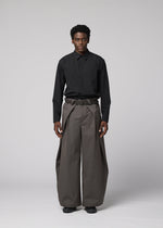A person stands facing forward against a gray background, wearing IM MEN's OVERLAP—black collared shirt tucked into high-waisted, wide natural-textured pants, belted at the waist and paired with black shoes.