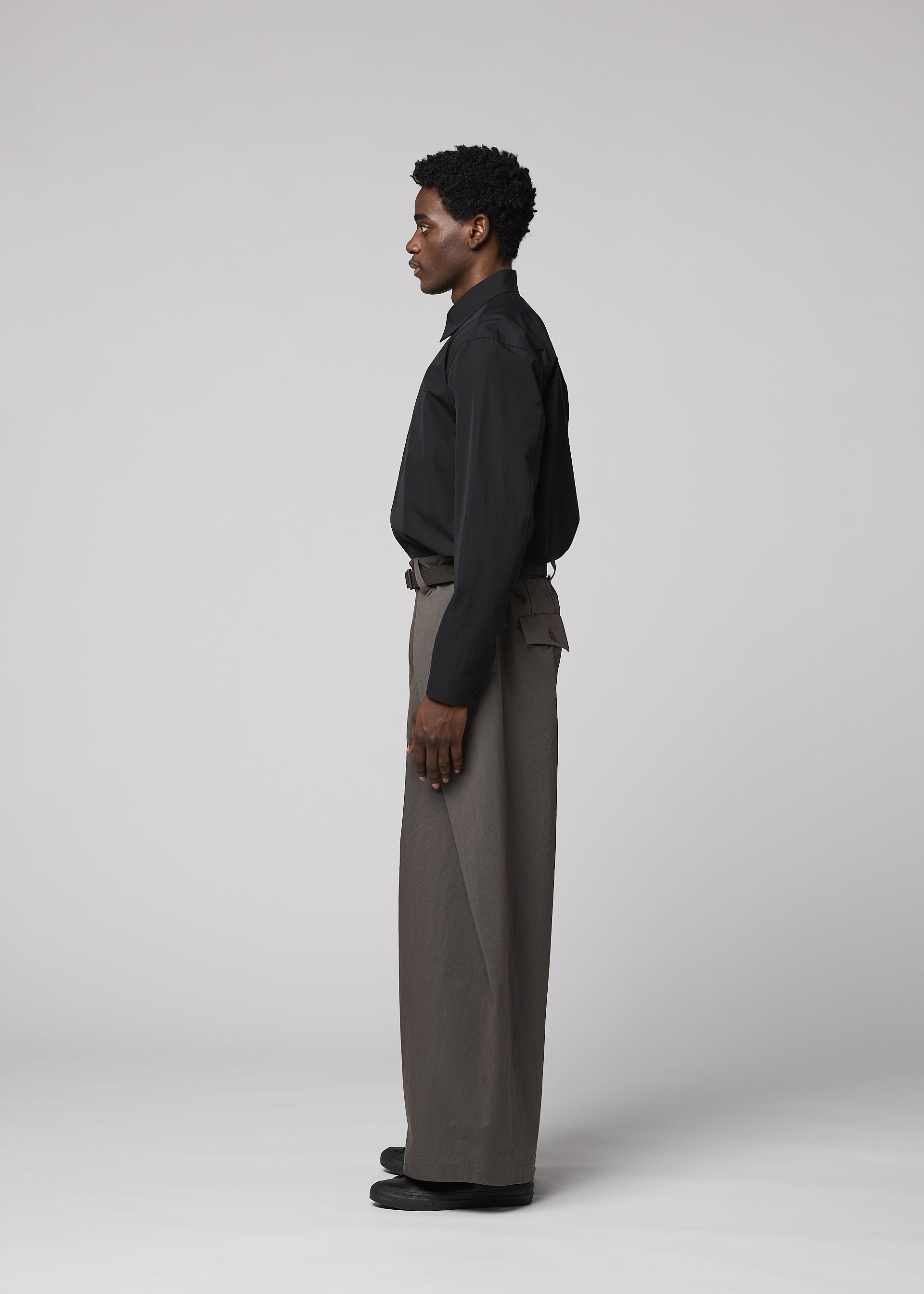 Zoomed view A man models IM MEN's OVERLAP collection, wearing a black button-up shirt and wide cotton-nylon blend pants, standing in profile against a plain light gray background with relaxed arms at his sides.