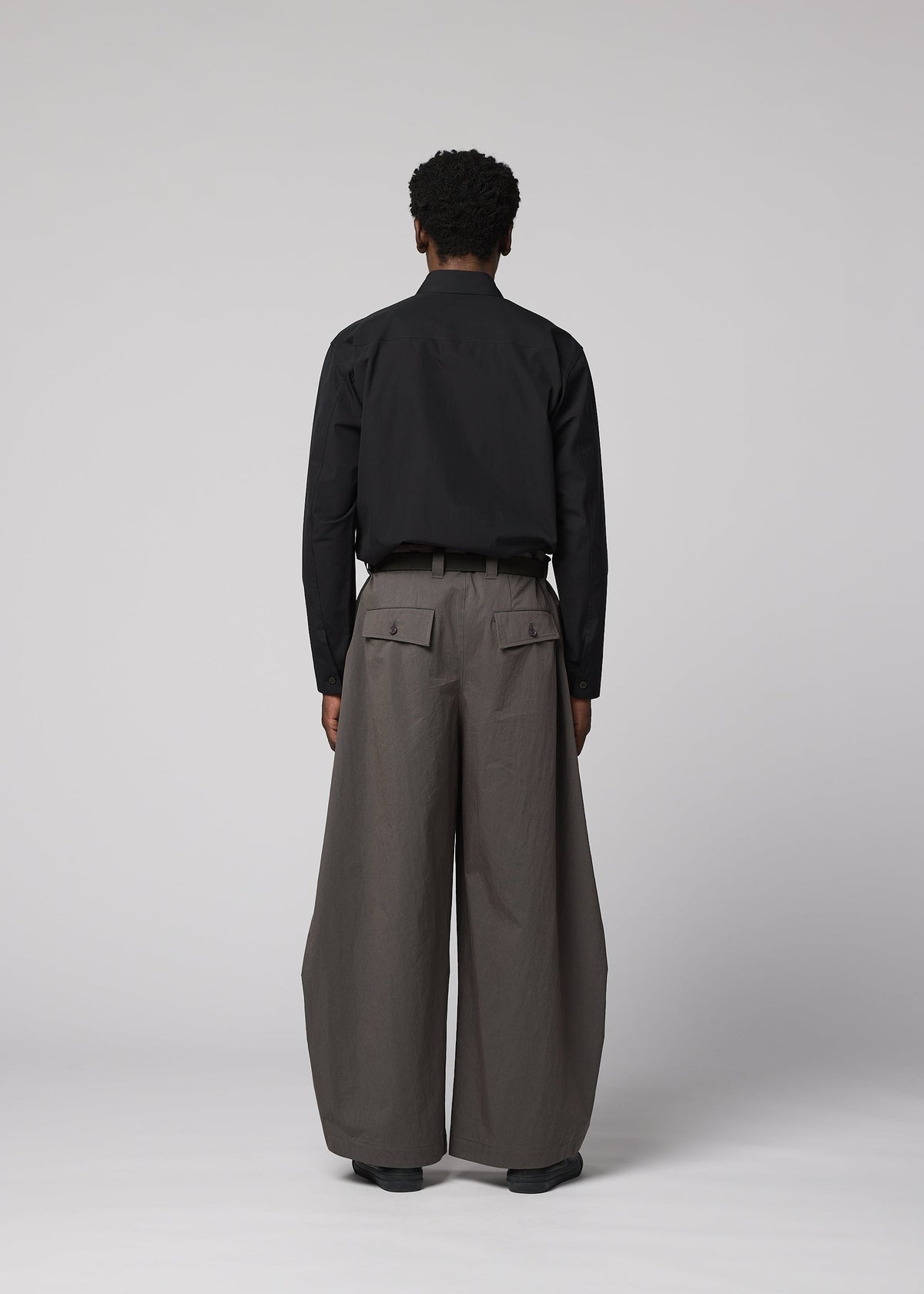 A person faces away, wearing IM MEN's OVERLAP: black long-sleeve shirt tucked into wide, high-waisted gray trousers with large pockets. The textured cotton-nylon blend pants stand out against a plain light gray background. view 7 of 8