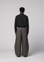 A person faces away, wearing IM MEN's OVERLAP: black long-sleeve shirt tucked into wide, high-waisted gray trousers with large pockets. The textured cotton-nylon blend pants stand out against a plain light gray background.