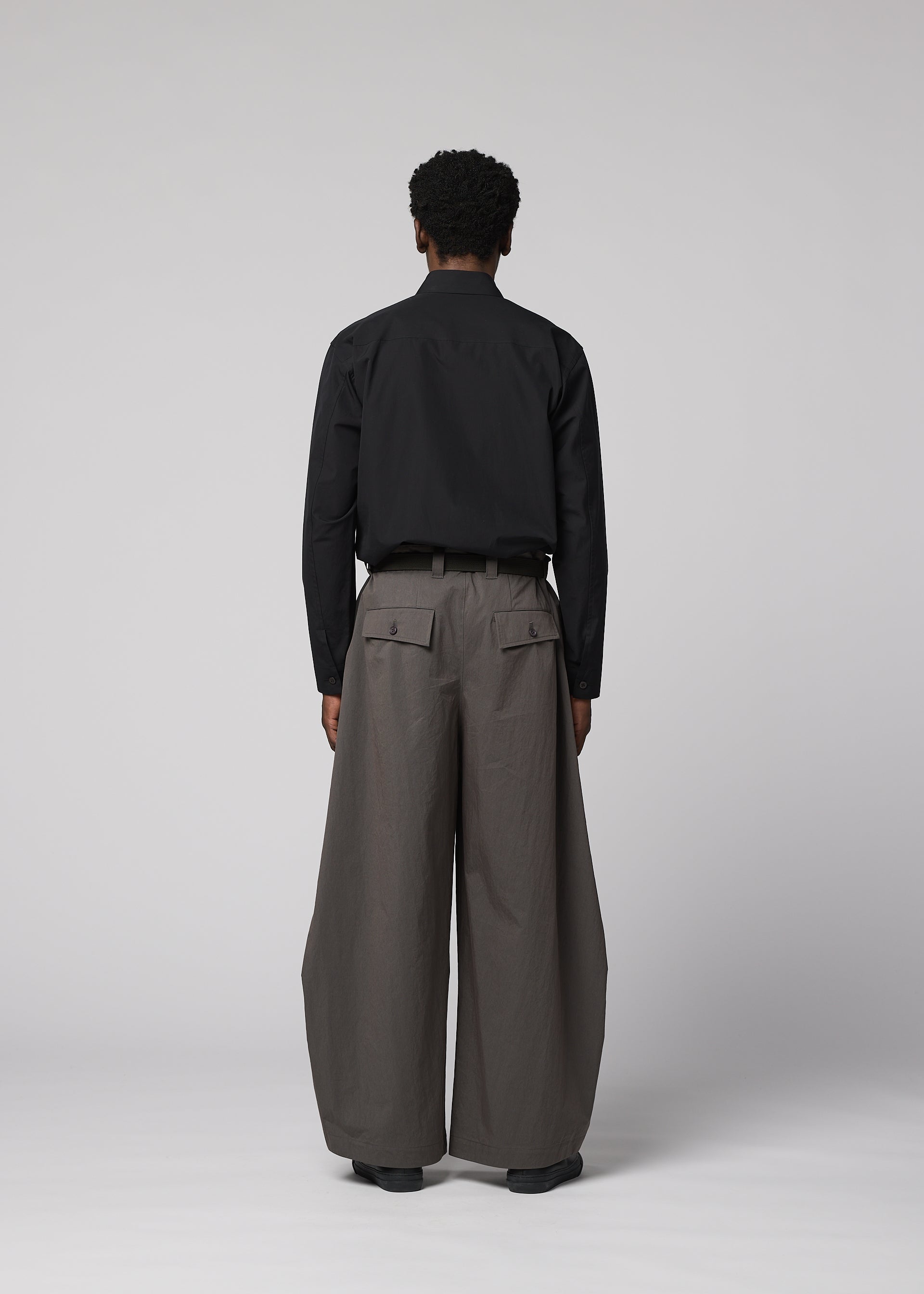 Zoomed view A person faces away, wearing IM MEN's OVERLAP: black long-sleeve shirt tucked into wide, high-waisted gray trousers with large pockets. The textured cotton-nylon blend pants stand out against a plain light gray background.