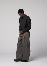 A man stands against a light gray background, wearing IM MEN’s OVERLAP black long-sleeve shirt and oversized wide pants in a natural texture, looking downward with his hands loosely clasped.