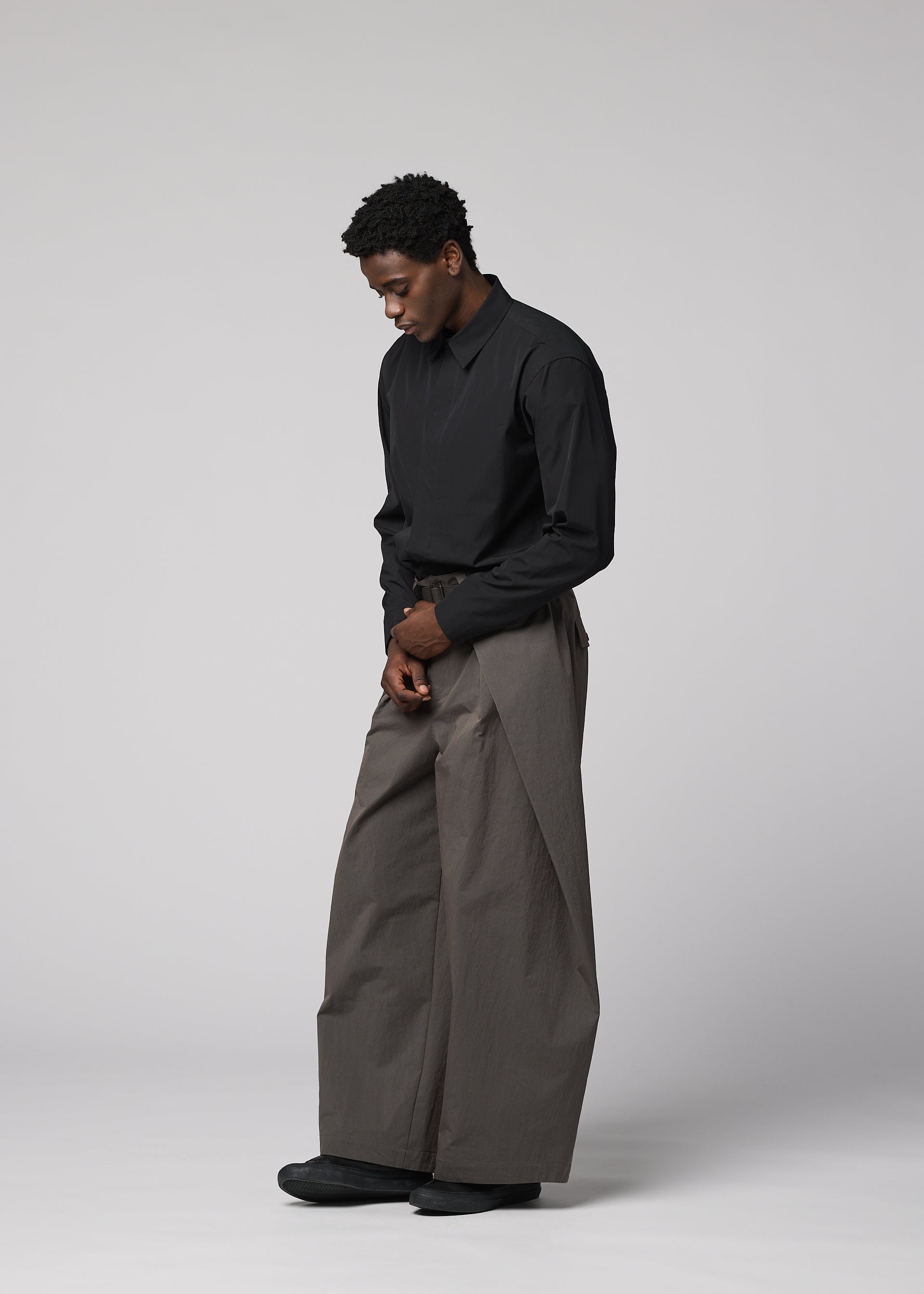 Zoomed view A man stands against a light gray background, wearing IM MEN’s OVERLAP black long-sleeve shirt and oversized wide pants in a natural texture, looking downward with his hands loosely clasped.