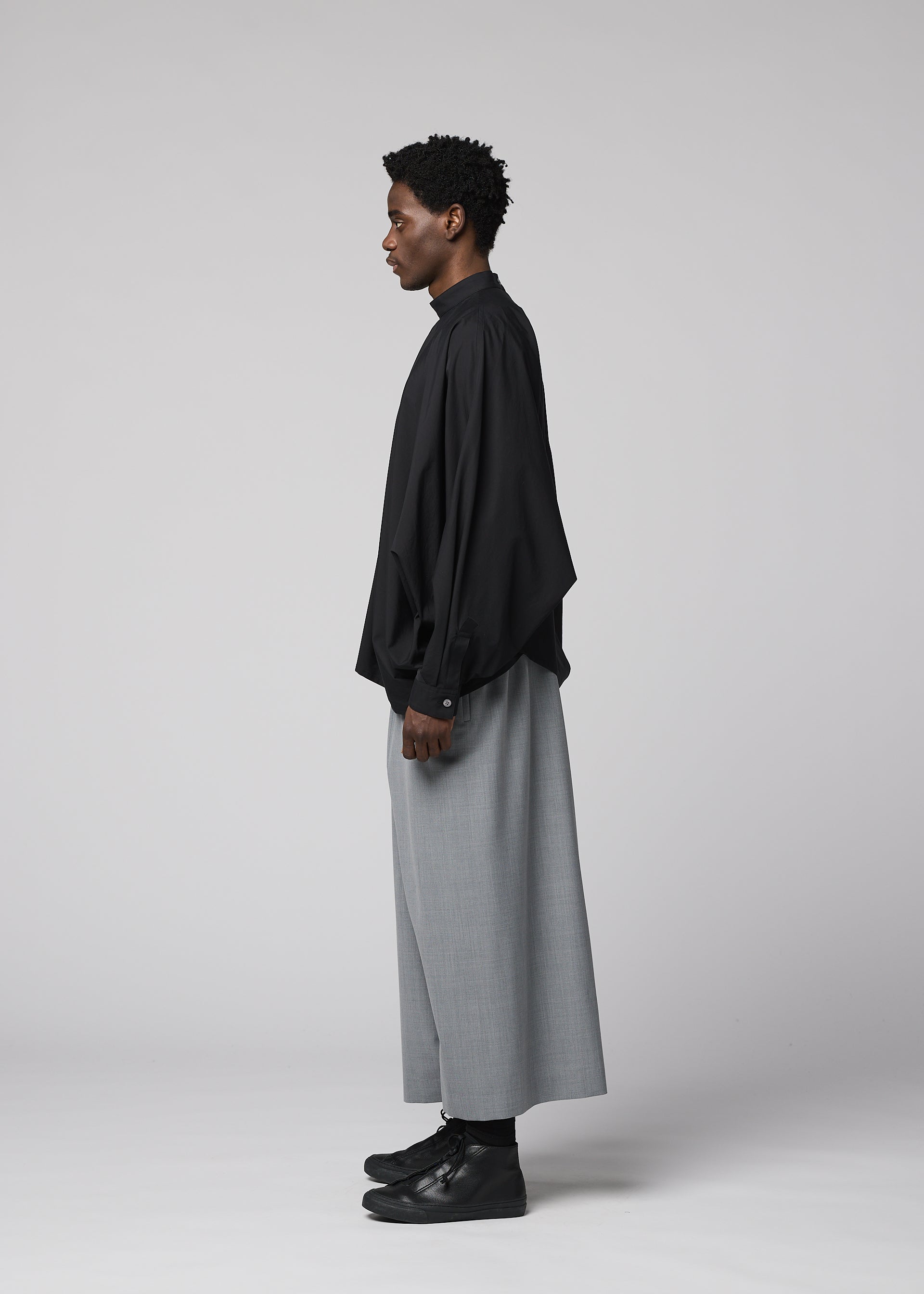 Zoomed view A person stands in profile against a plain backdrop, wearing IM MEN’s SUAVE loose black shirt and wide wool blend sarouel pants with black shoes—a minimalist, modern look with a relaxed silhouette.
