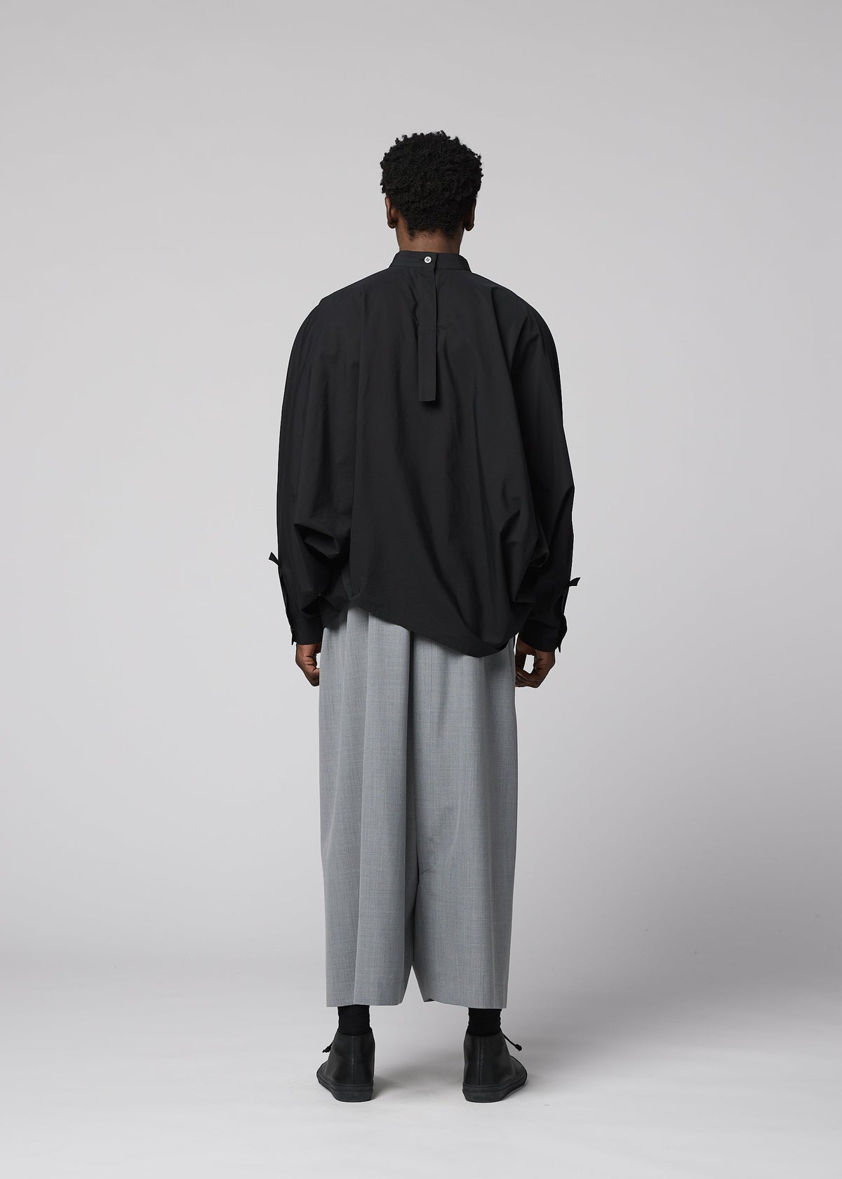 A person with short curly hair wears the IM MEN SUAVE loose black long-sleeve shirt with buttoned cuffs and wide wool blend sarouel pants, paired with black shoes, standing against a plain light gray background. view 8 of 9