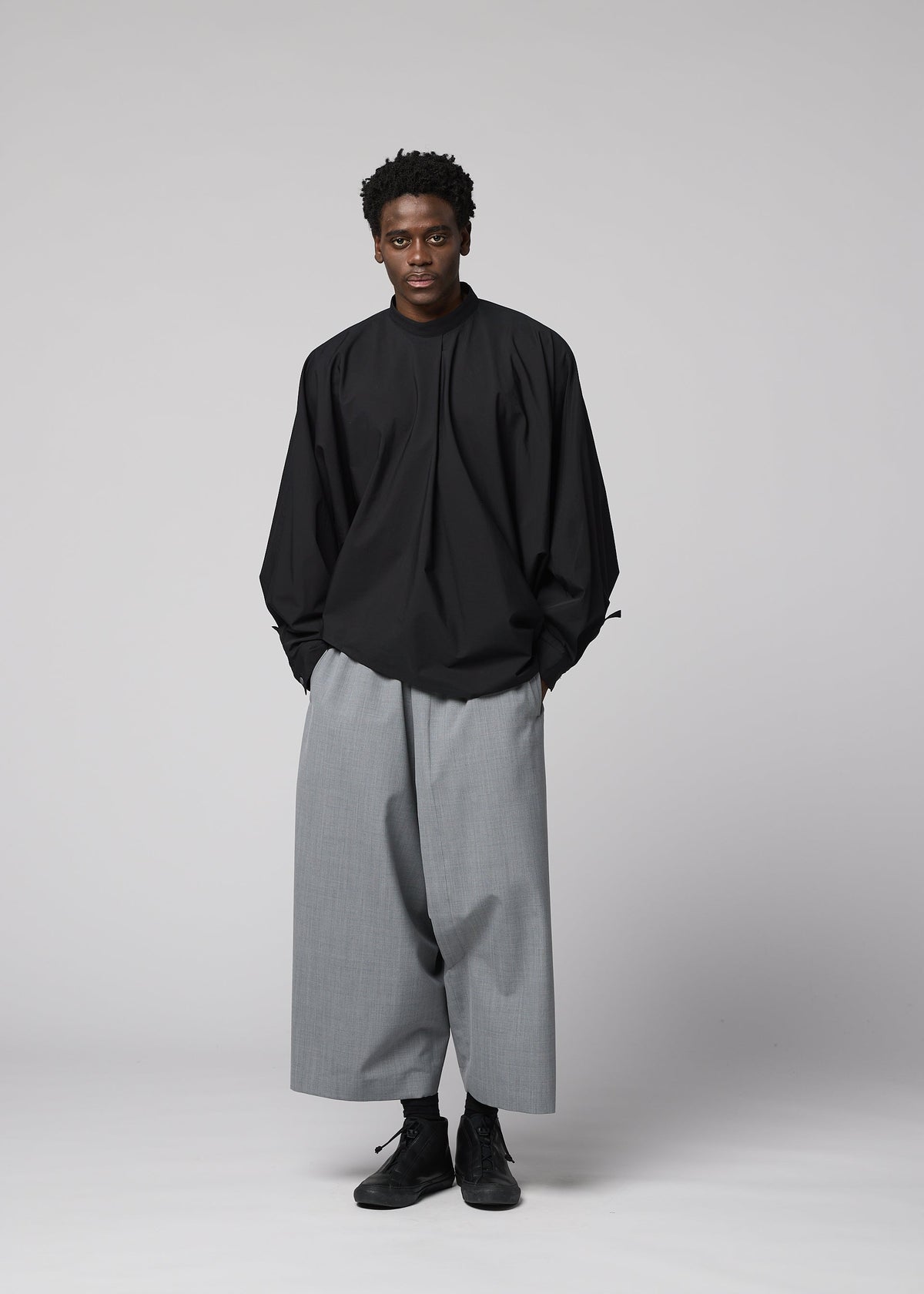 A person poses against a plain light gray background, wearing the IM MEN SUAVE loose-fitting black long-sleeved top and wide wool blend sarouel pants, hands in pockets and a neutral expression complementing the relaxed outfit. view 9 of 9