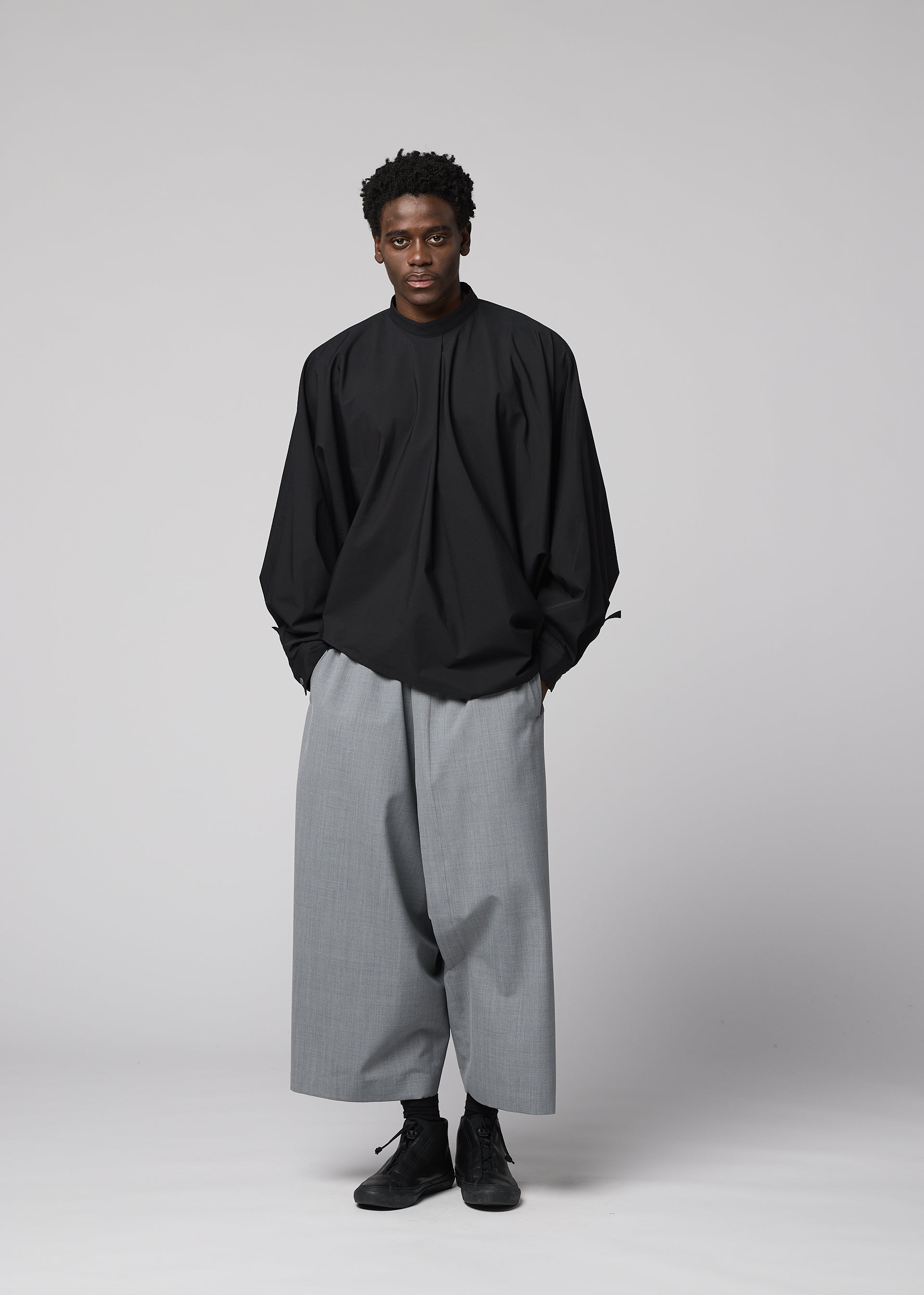 Zoomed view A person poses against a plain light gray background, wearing the IM MEN SUAVE loose-fitting black long-sleeved top and wide wool blend sarouel pants, hands in pockets and a neutral expression complementing the relaxed outfit.