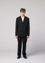 A person with short, tousled hair stands facing forward in IM MEN’s SUAVE: a loose-fitting black suit of lightweight wool pants, white shirt, and black shoes, set against a plain light gray background.