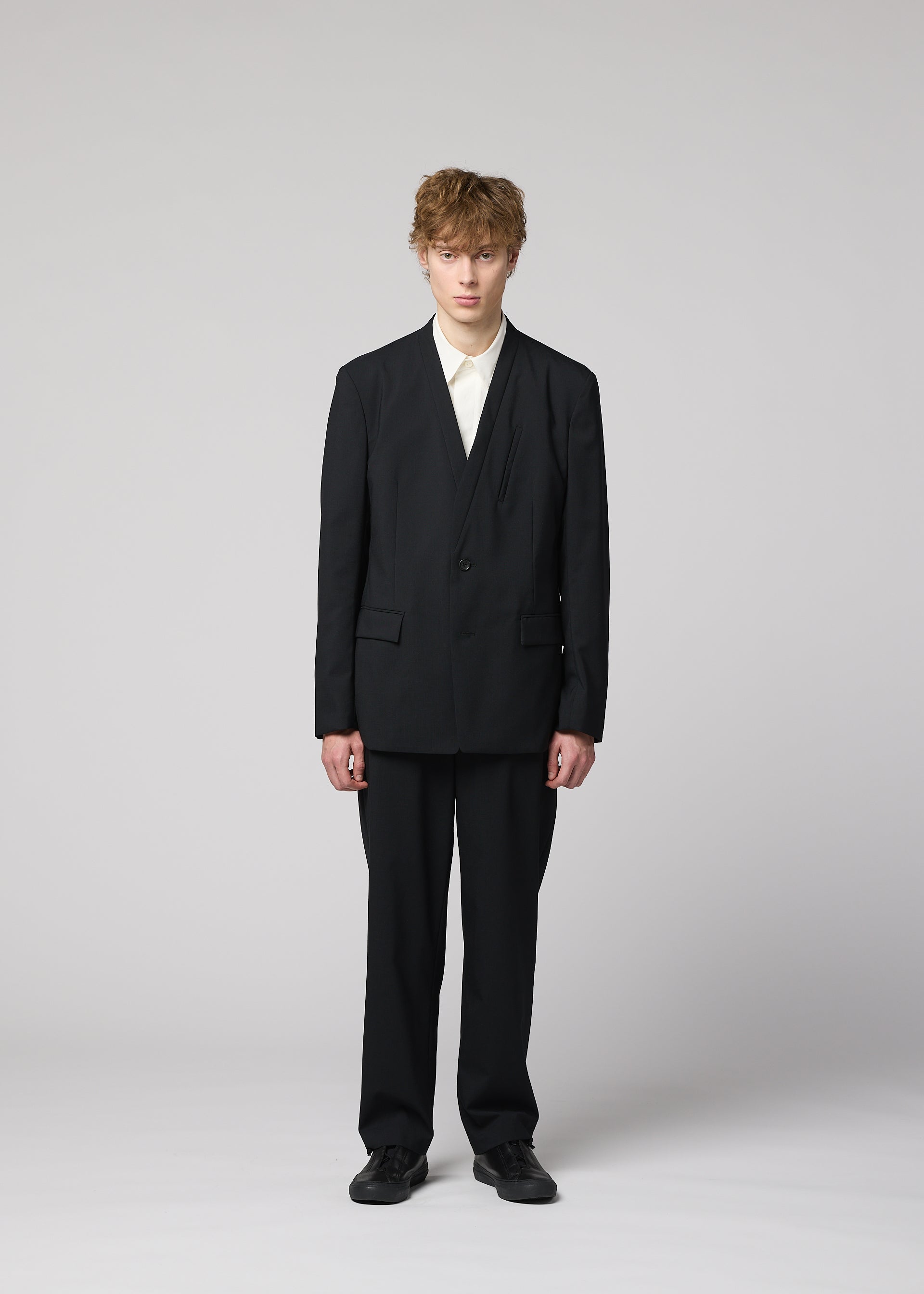 Zoomed view A person with short, tousled hair stands facing forward in IM MEN’s SUAVE: a loose-fitting black suit of lightweight wool pants, white shirt, and black shoes, set against a plain light gray background.