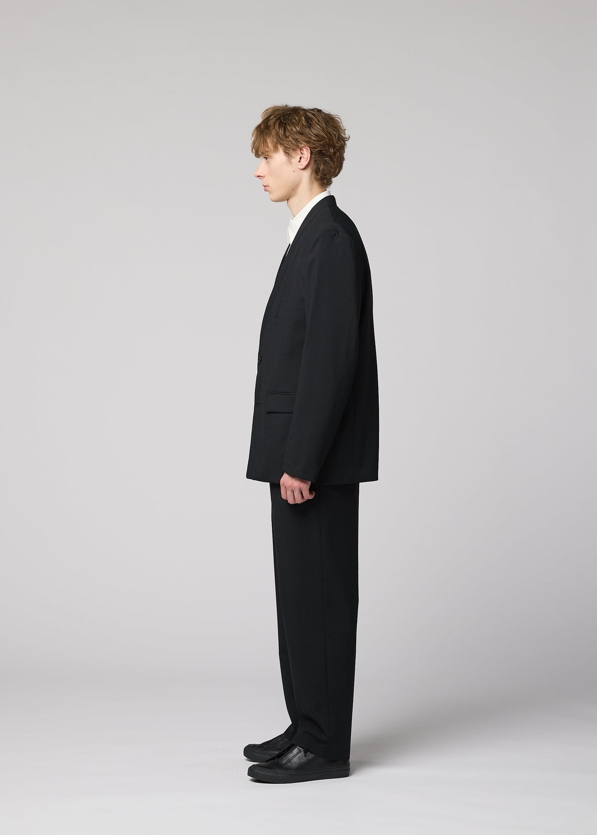 A young person with curly hair stands in profile against a light gray background, wearing the SUAVE black suit with breathable straight pants, white shirt, and black shoes from IM MEN. view 7 of 9