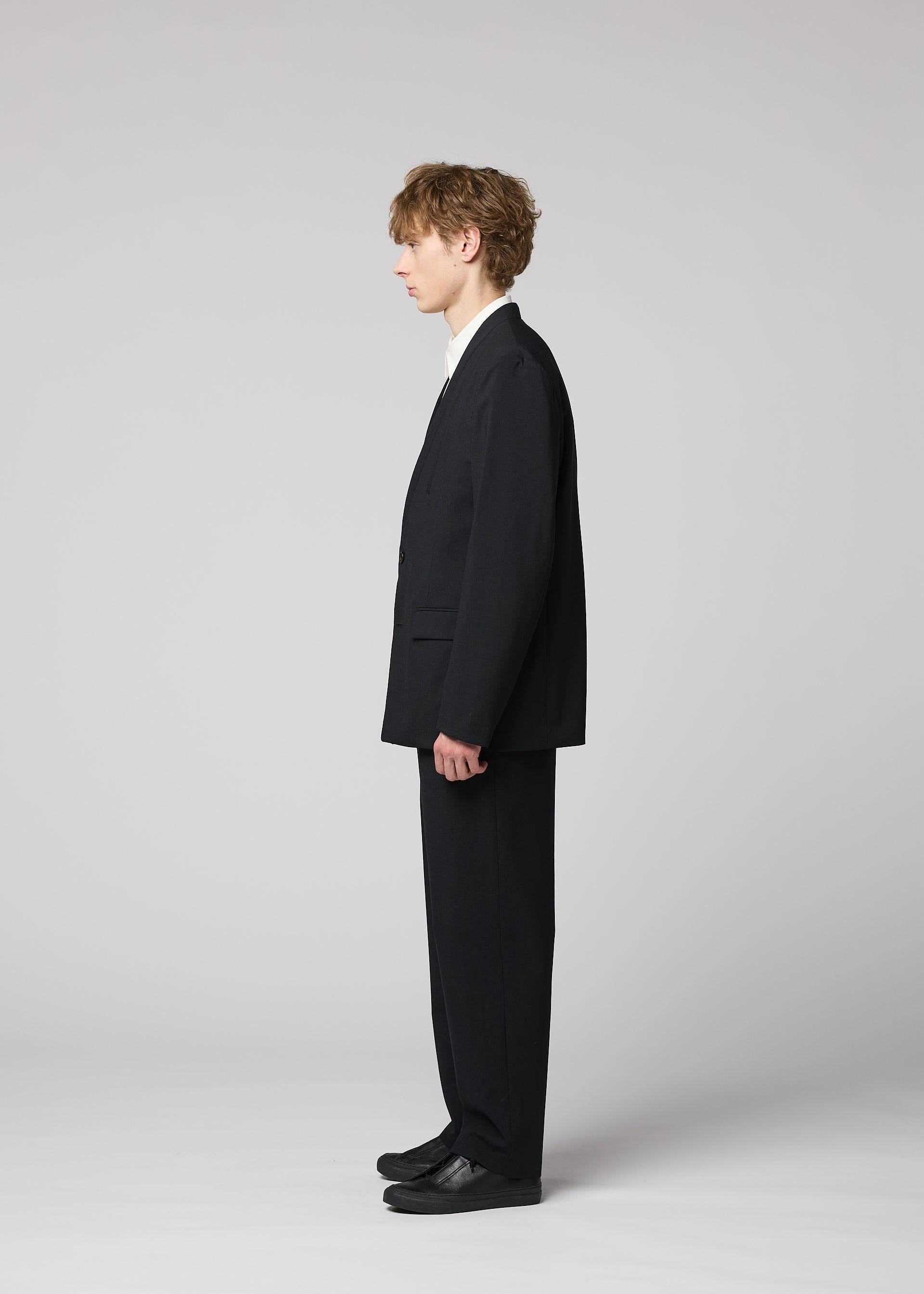 Zoomed view A young person with curly hair stands in profile against a light gray background, wearing the SUAVE black suit with breathable straight pants, white shirt, and black shoes from IM MEN.