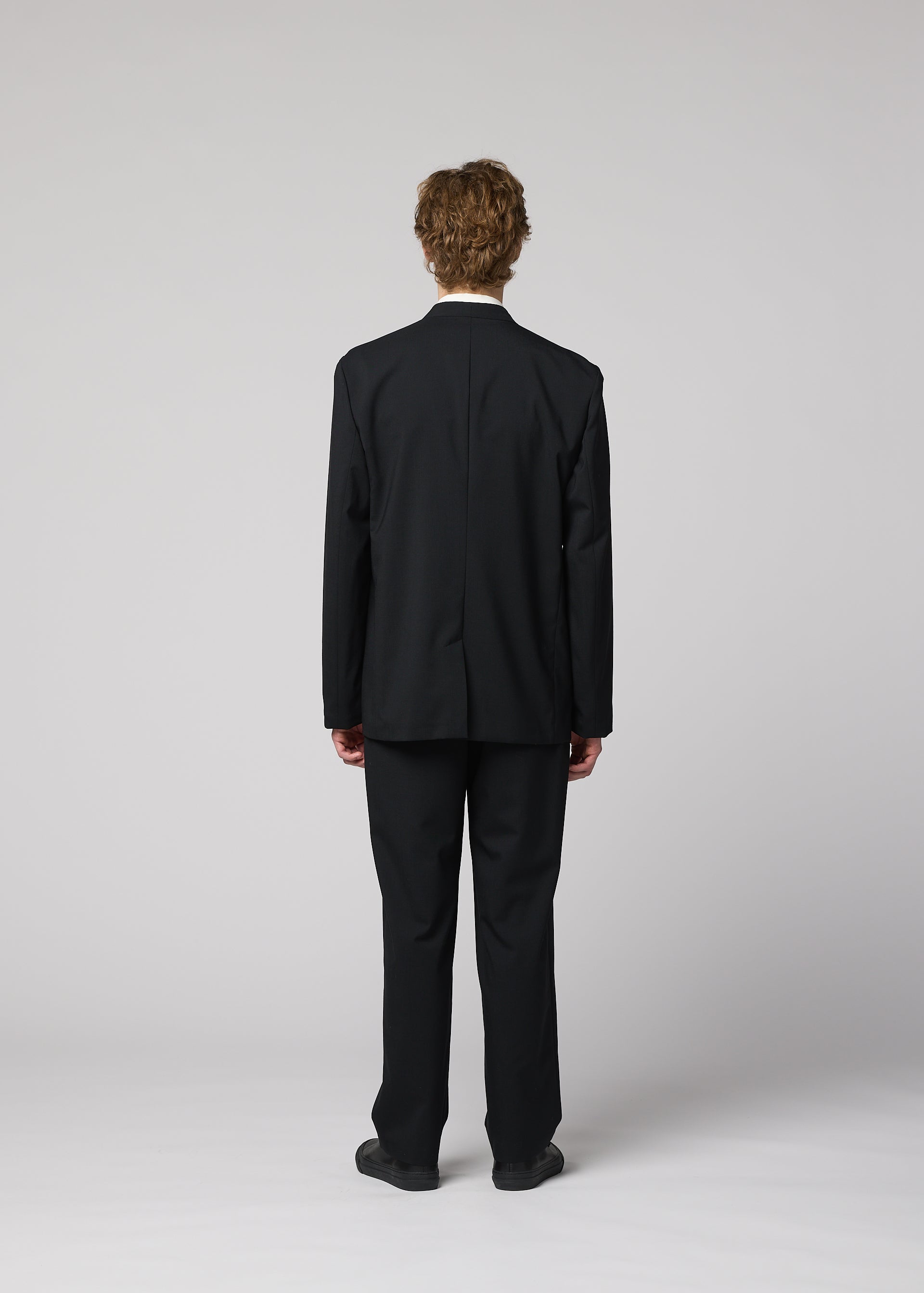 Zoomed view A person with curly hair, seen from behind, wears the IM MEN SUAVE black suit with lightweight wool pants and black shoes against a plain light gray background.