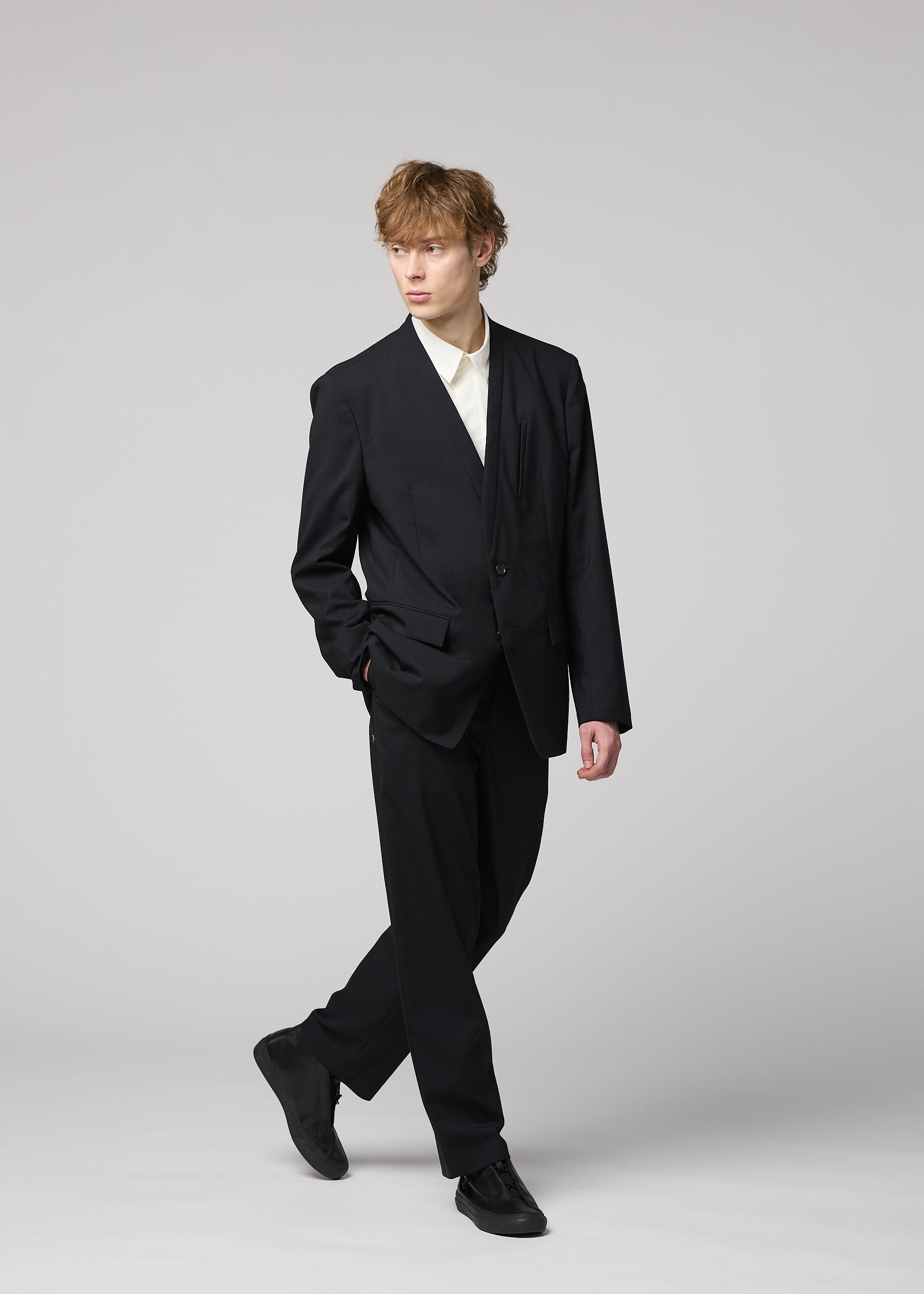 Zoomed view A young man with wavy light brown hair wears the SUAVE black suit and breathable straight pants from IM MEN over a white shirt. He stands against a light gray background, one hand in his pocket, looking to the side.
