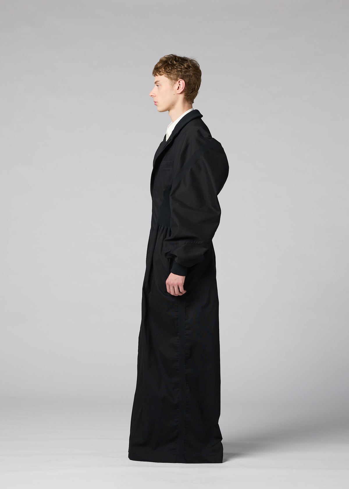 A person stands in profile against a plain gray background, wearing “CLAY,” a long, oversized black coat with puffy sleeves crafted from sculptural fabric, from the IM MEN Autumn Winter Collection. view 8 of 9