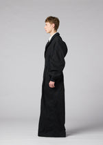 A person stands in profile against a plain gray background, wearing “CLAY,” a long, oversized black coat with puffy sleeves crafted from sculptural fabric, from the IM MEN Autumn Winter Collection.