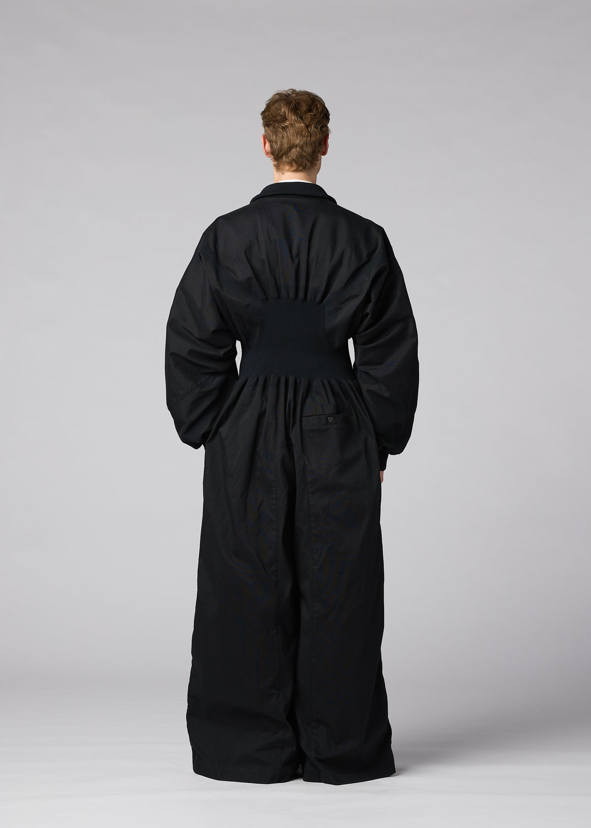 A person stands facing away from the camera, wearing CLAY—a loose-fitting black rib knit jumpsuit with a cinched waist from IM MEN—set against a plain gray background. view 9 of 9
