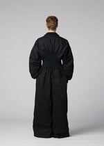 A person stands facing away from the camera, wearing CLAY—a loose-fitting black rib knit jumpsuit with a cinched waist from IM MEN—set against a plain gray background.