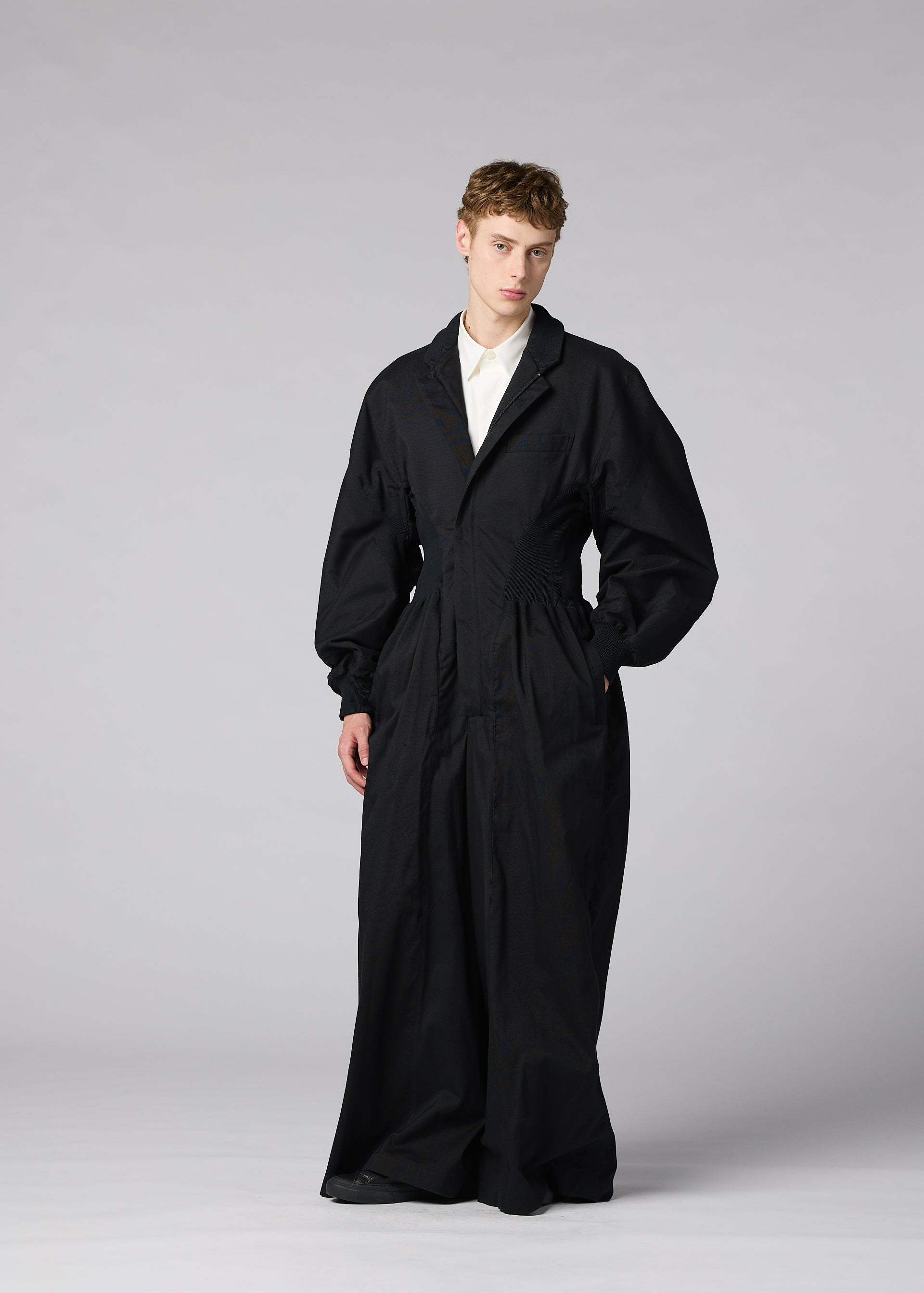 Zoomed view A person with short, curly hair wears the long, oversized black “CLAY” coat from IM MEN over a white collared shirt, standing against a plain gray background.
