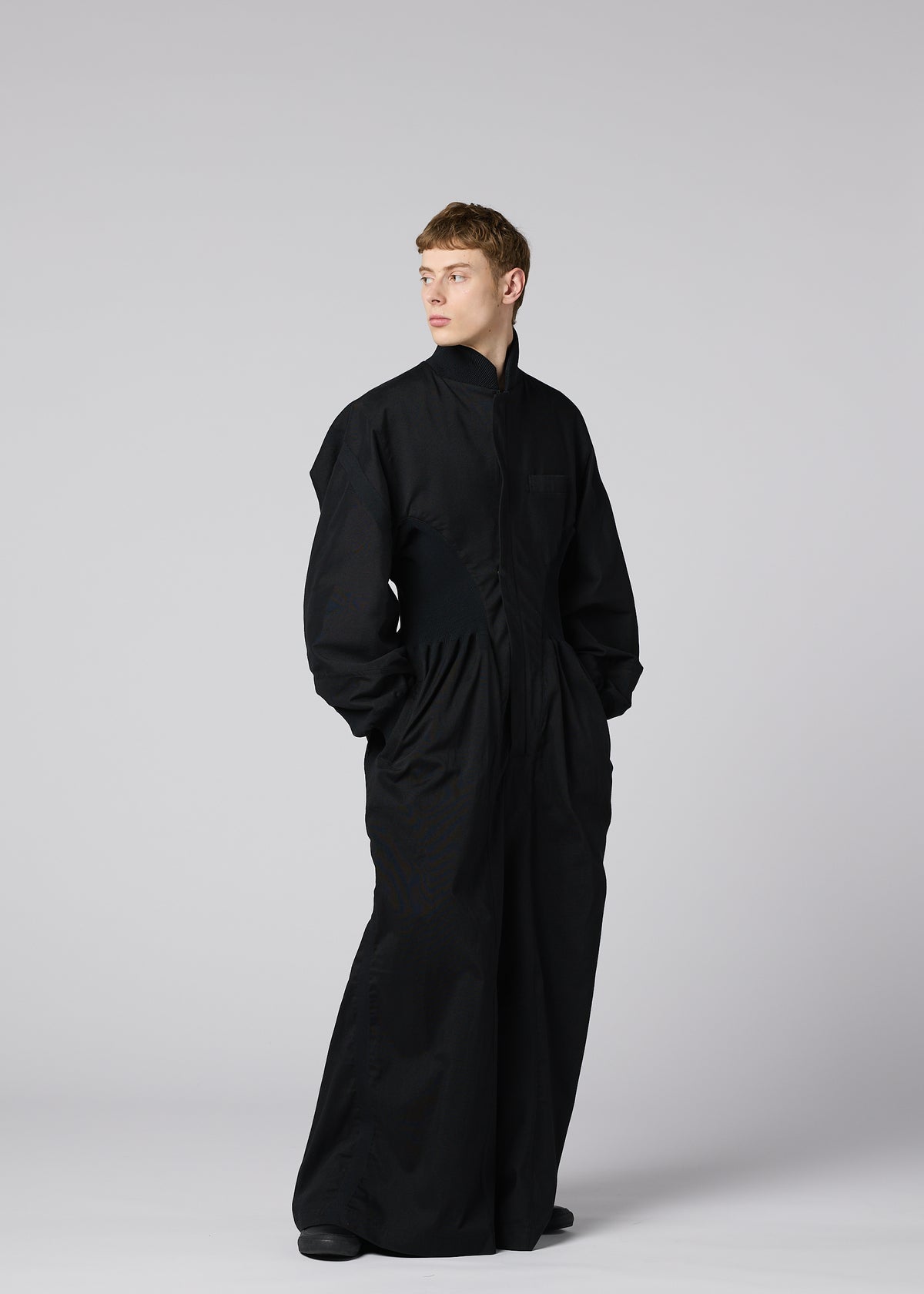 Zoomed view A person stands against a light gray background wearing the CLAY black rib knit jumpsuit from IM MEN’s Autumn Winter Collection. The relaxed-fit jumpsuit features long sleeves, wide legs, and pockets, and the wearer looks off to the side.