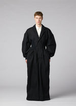 A model stands in a studio wearing IM MEN’s CLAY—an oversized, long black coat crafted from sculptural fabric—over a white collared shirt. The background is plain gray and the individual faces forward with arms at their sides.