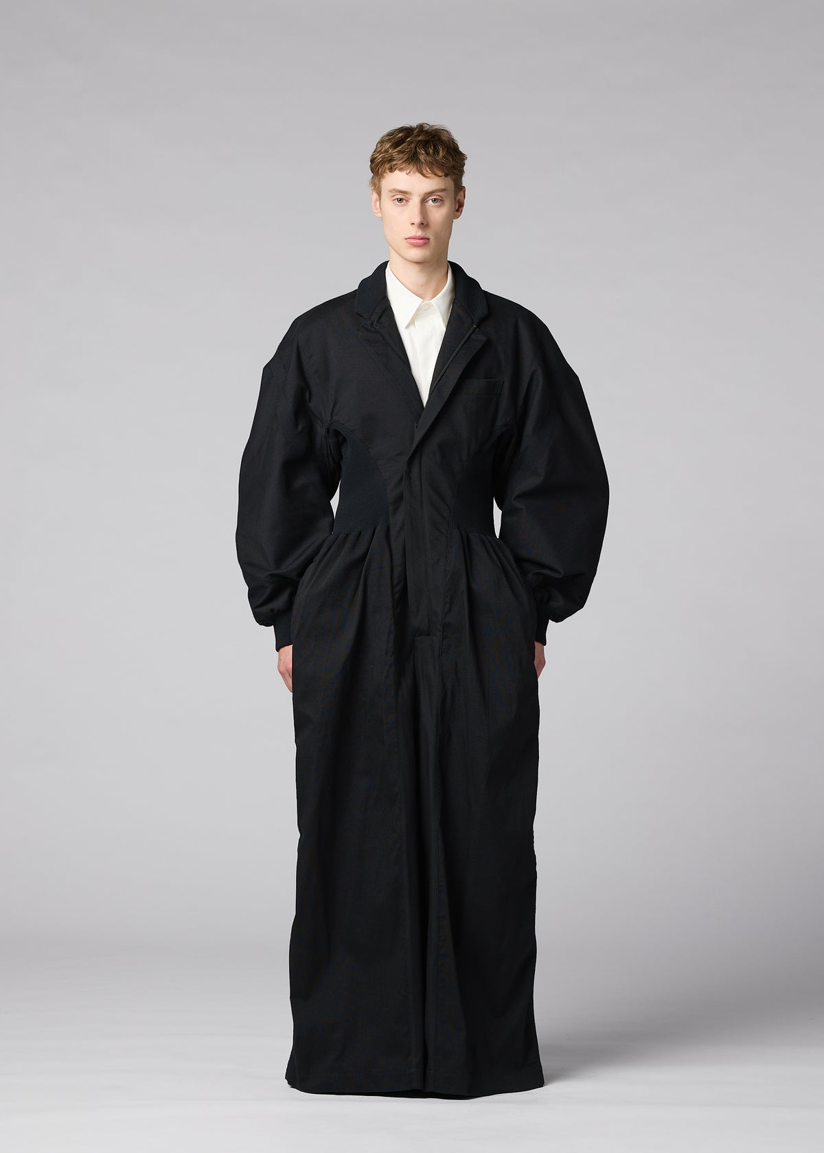Zoomed view A model stands in a studio wearing IM MEN’s CLAY—an oversized, long black coat crafted from sculptural fabric—over a white collared shirt. The background is plain gray and the individual faces forward with arms at their sides.