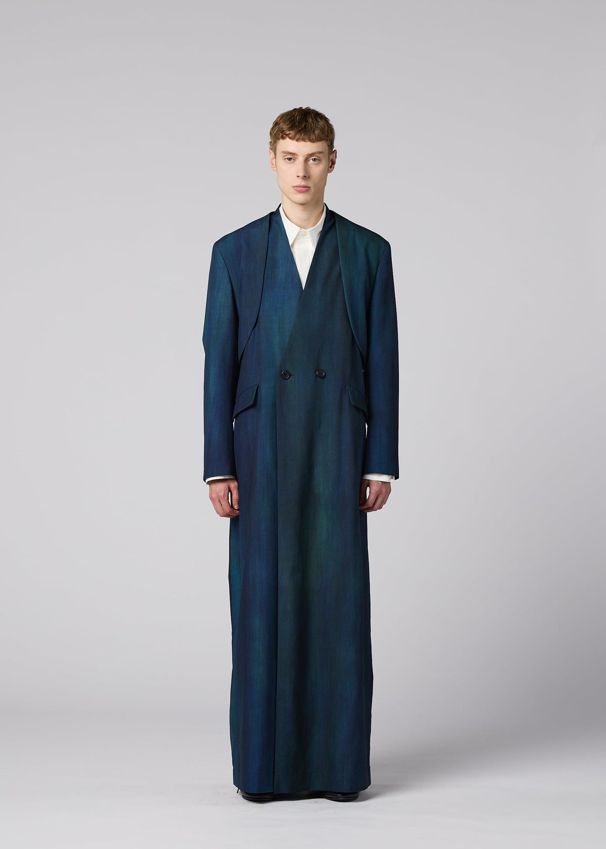 A person stands facing forward in an oversized, floor-length blue GRADATION WOOL coat from IM MEN over a white shirt, against a plain gray background—part of the Autumn Winter 2026/27 collection. view 9 of 13