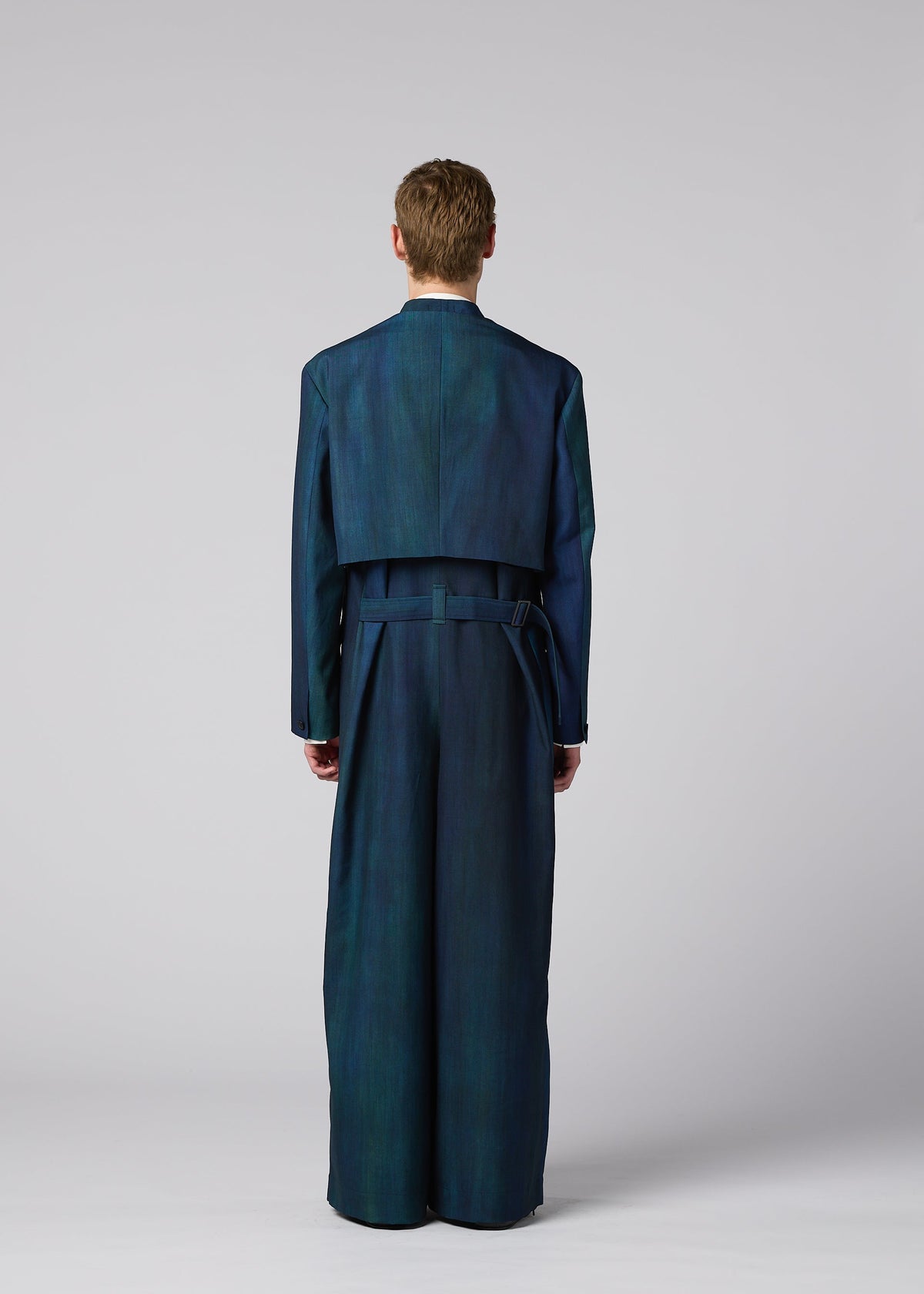 A person stands facing away in IM MEN’s GRADATION WOOL blue-green suit with cropped jacket and wide-leg trousers, set against a plain light gray background. Autumn Winter 2026/27. view 11 of 13