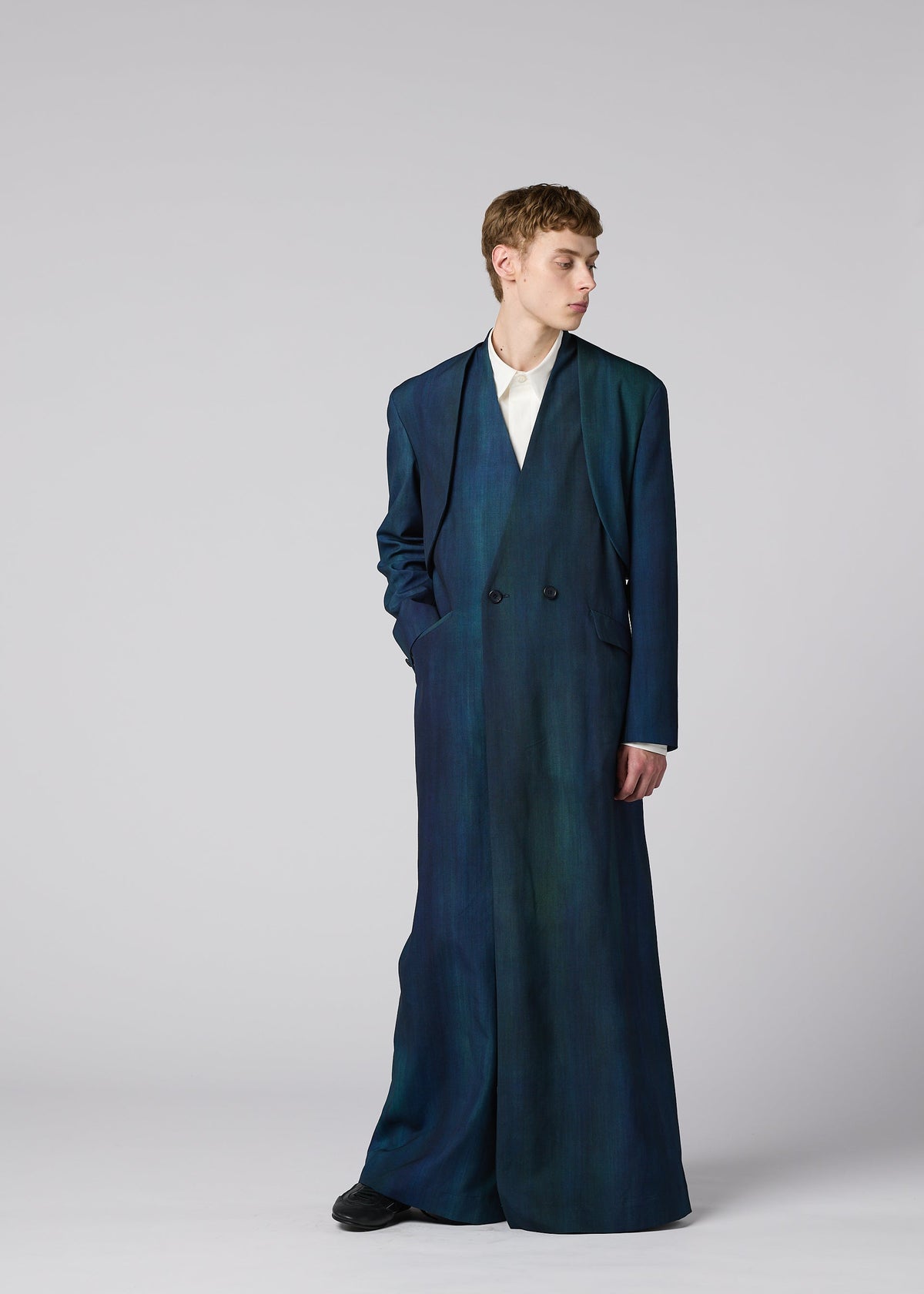 Zoomed view A person stands against a plain background wearing the IM MEN GRADATION WOOL long dark blue coat over a white top, hands in pockets, looking to the side.