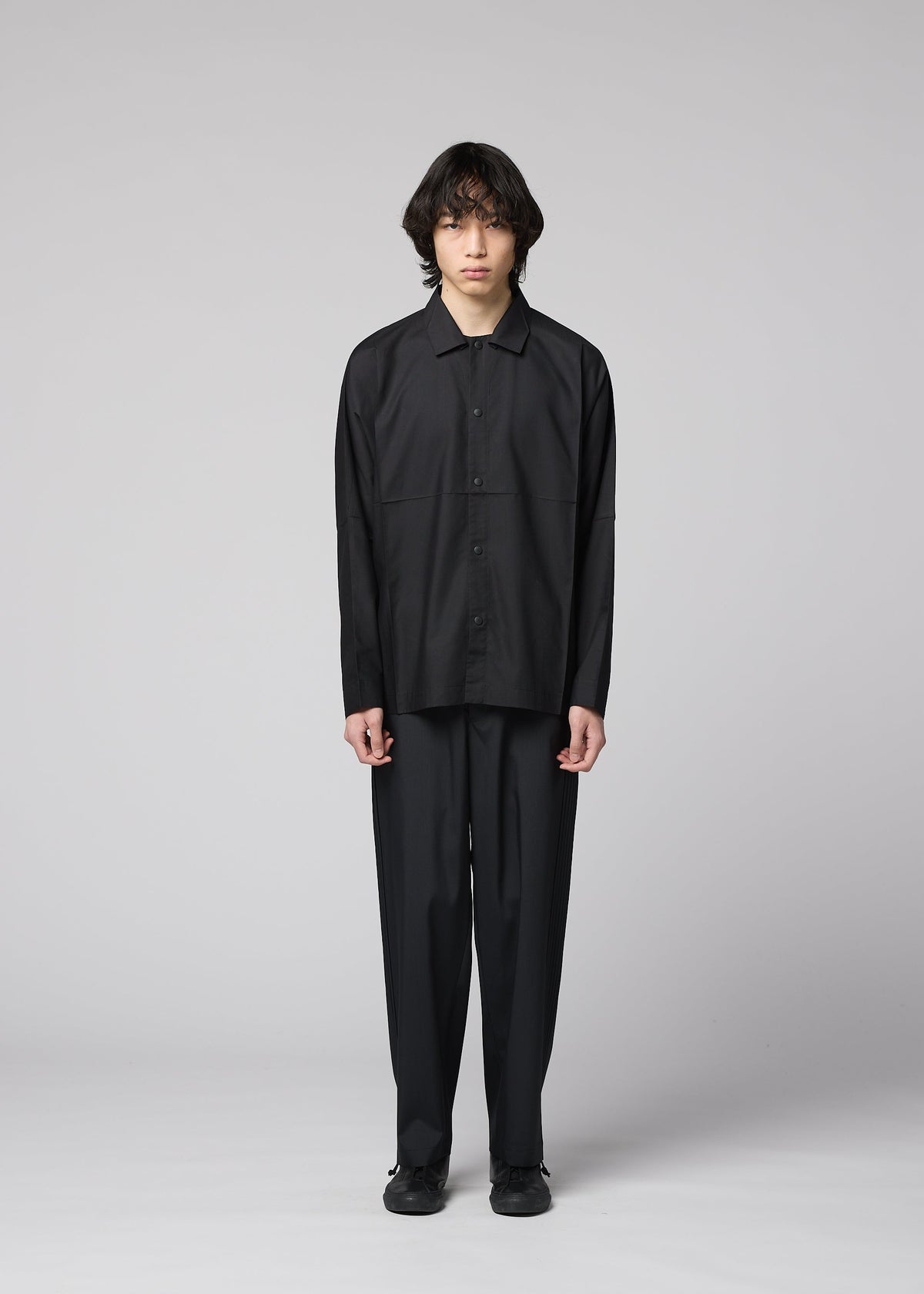 A person with medium-length dark hair faces forward against a plain gray background, wearing the IM MEN COMPACT SHIRT BLACK along with wide black pants and black shoes. view 8 of 11