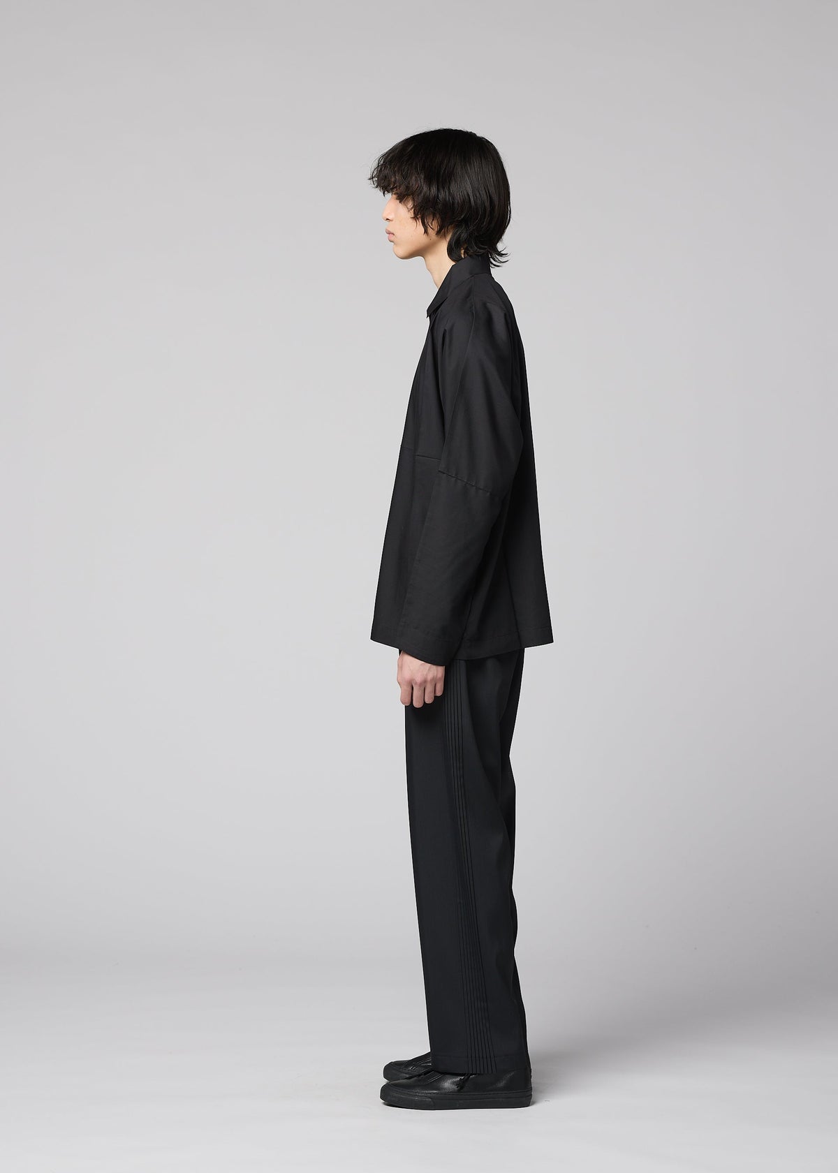 A person with medium-length dark hair stands in profile against a light gray background, wearing the IM MEN COMPACT SHIRT BLACK, complemented by matching black pants and black shoes. view 9 of 11