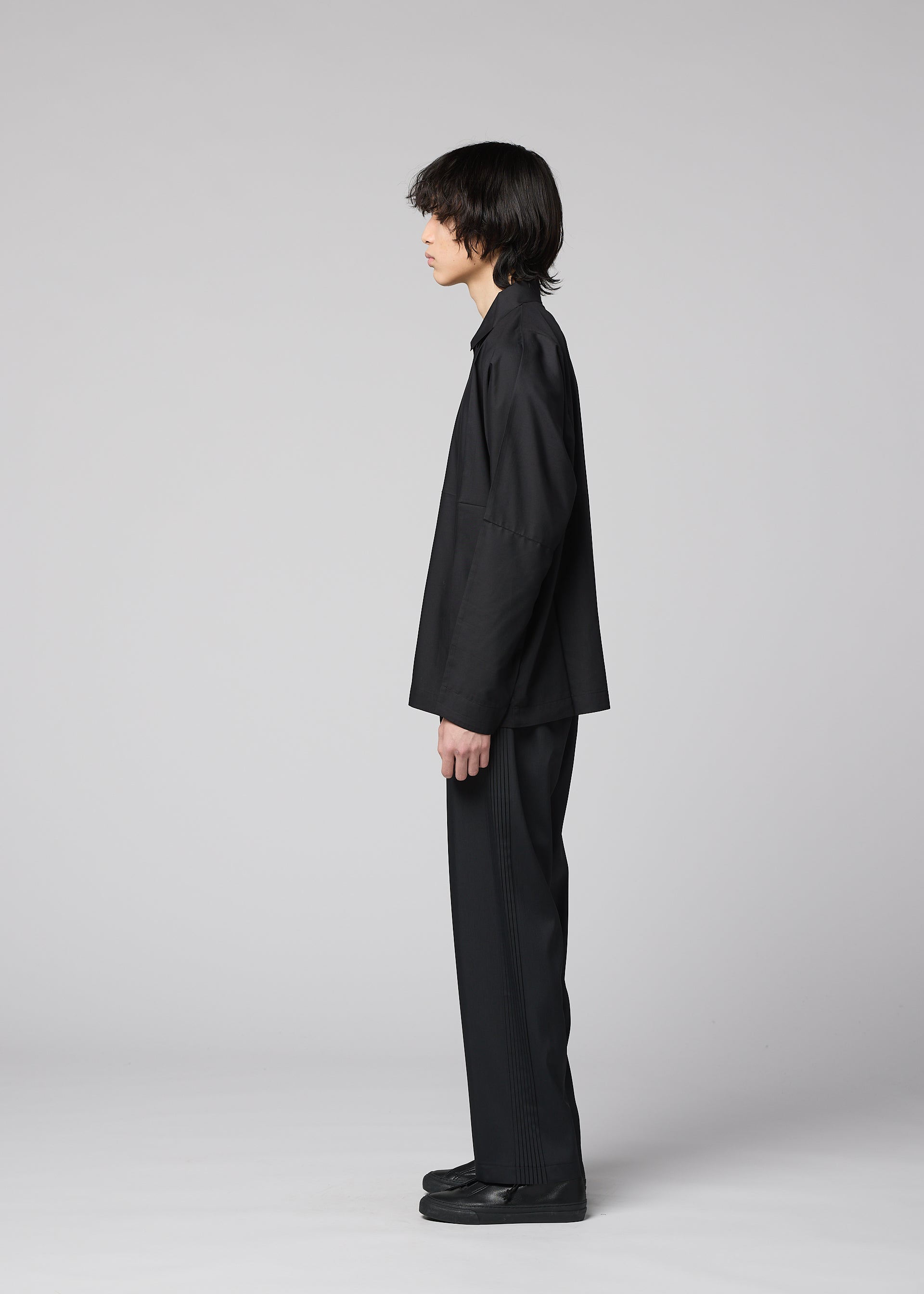 Zoomed view A person with medium-length dark hair stands in profile against a light gray background, wearing the IM MEN COMPACT SHIRT BLACK, complemented by matching black pants and black shoes.
