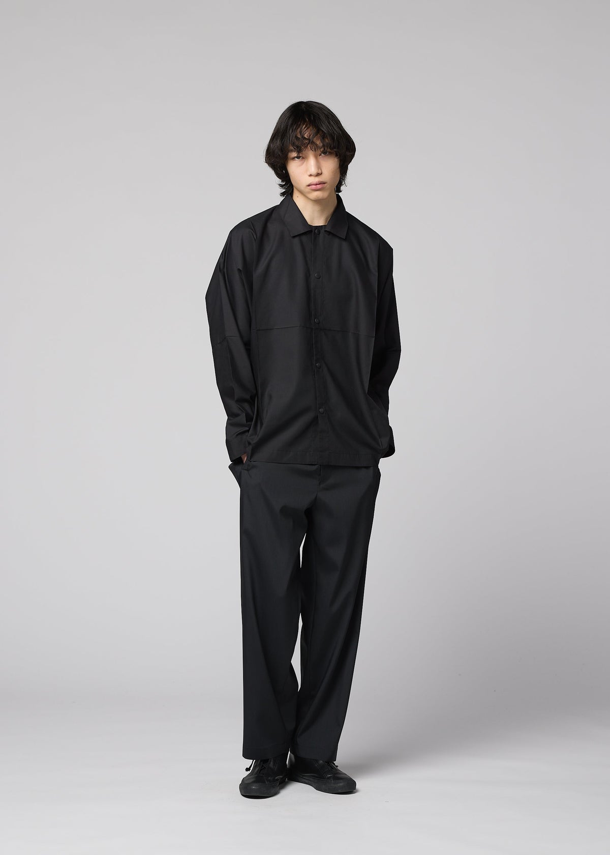 A person with medium-length dark hair stands against a plain light gray background, wearing the IM MEN COMPACT SHIRT BLACK, matching black pants and shoes, with hands in pockets and a neutral expression. view 11 of 11