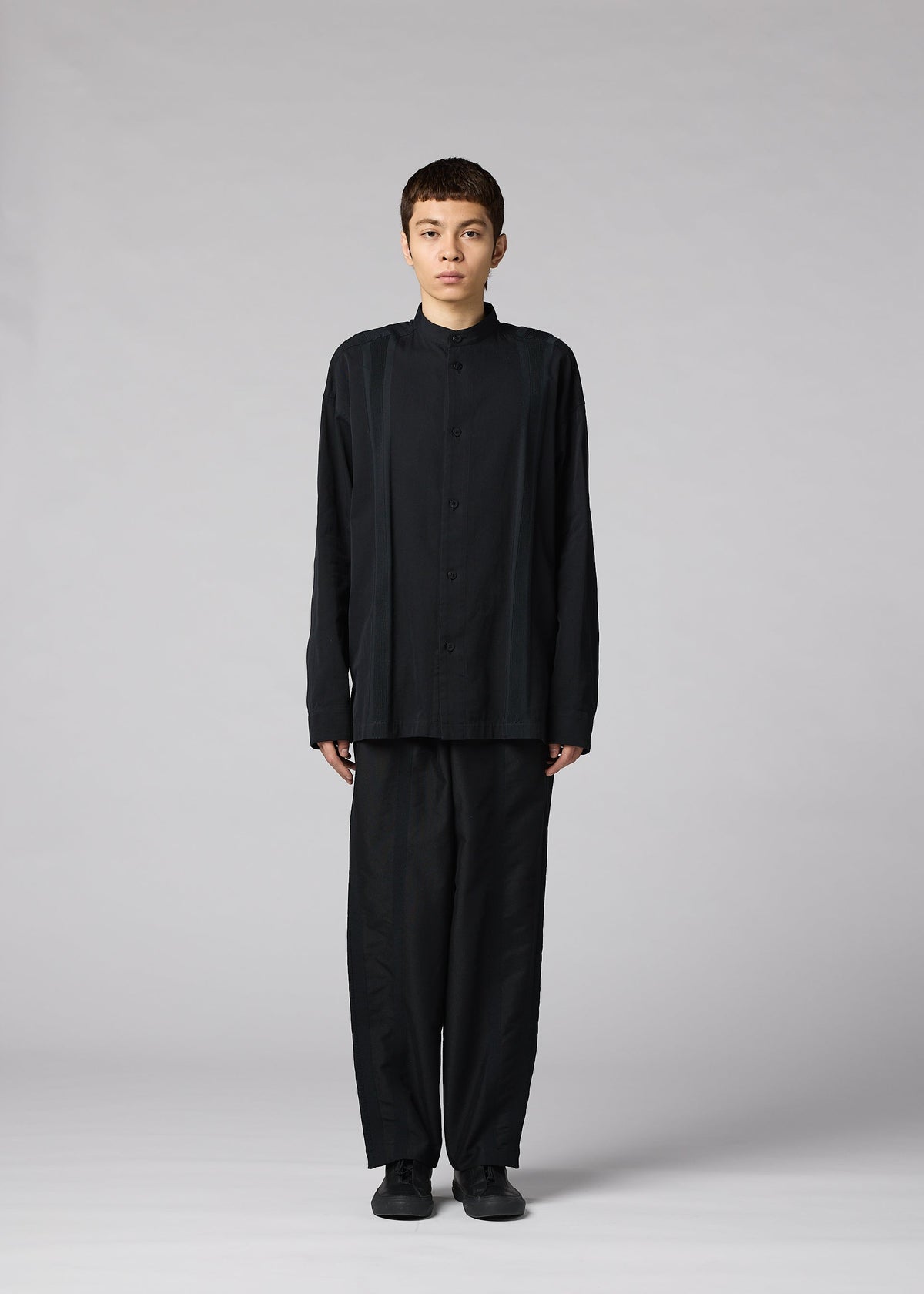 A person with short hair stands facing forward, dressed in the IM MEN CLAY SHIRT—a loose black cotton stand-up collar shirt—paired with wide black pants and shoes, set against a plain light gray background. view 5 of 8