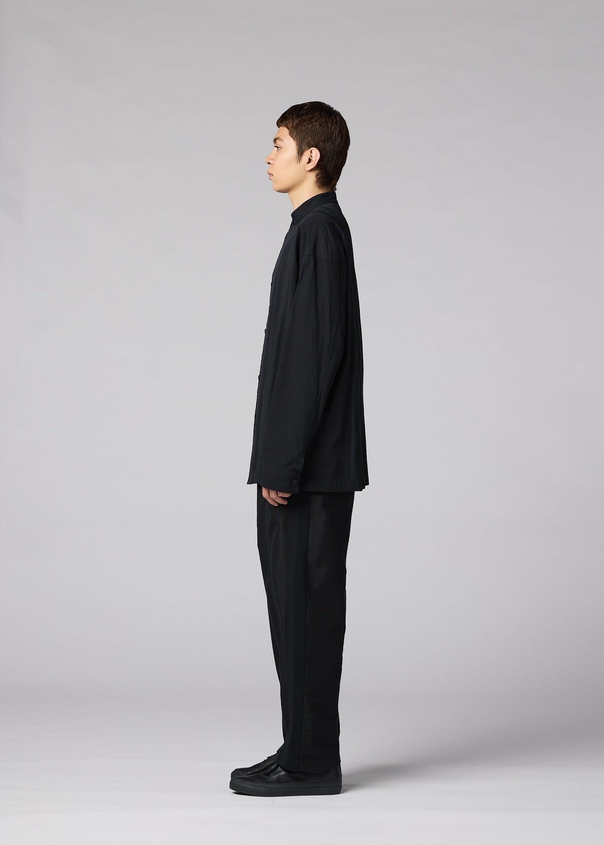 A person stands in profile facing left, wearing the IM MEN CLAY SHIRT—a loose-fitting black cotton stand-up collar shirt—and black cotton pants with black shoes, set against a plain gray background. view 6 of 8