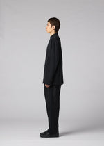 A person stands in profile facing left, wearing the IM MEN CLAY SHIRT—a loose-fitting black cotton stand-up collar shirt—and black cotton pants with black shoes, set against a plain gray background.