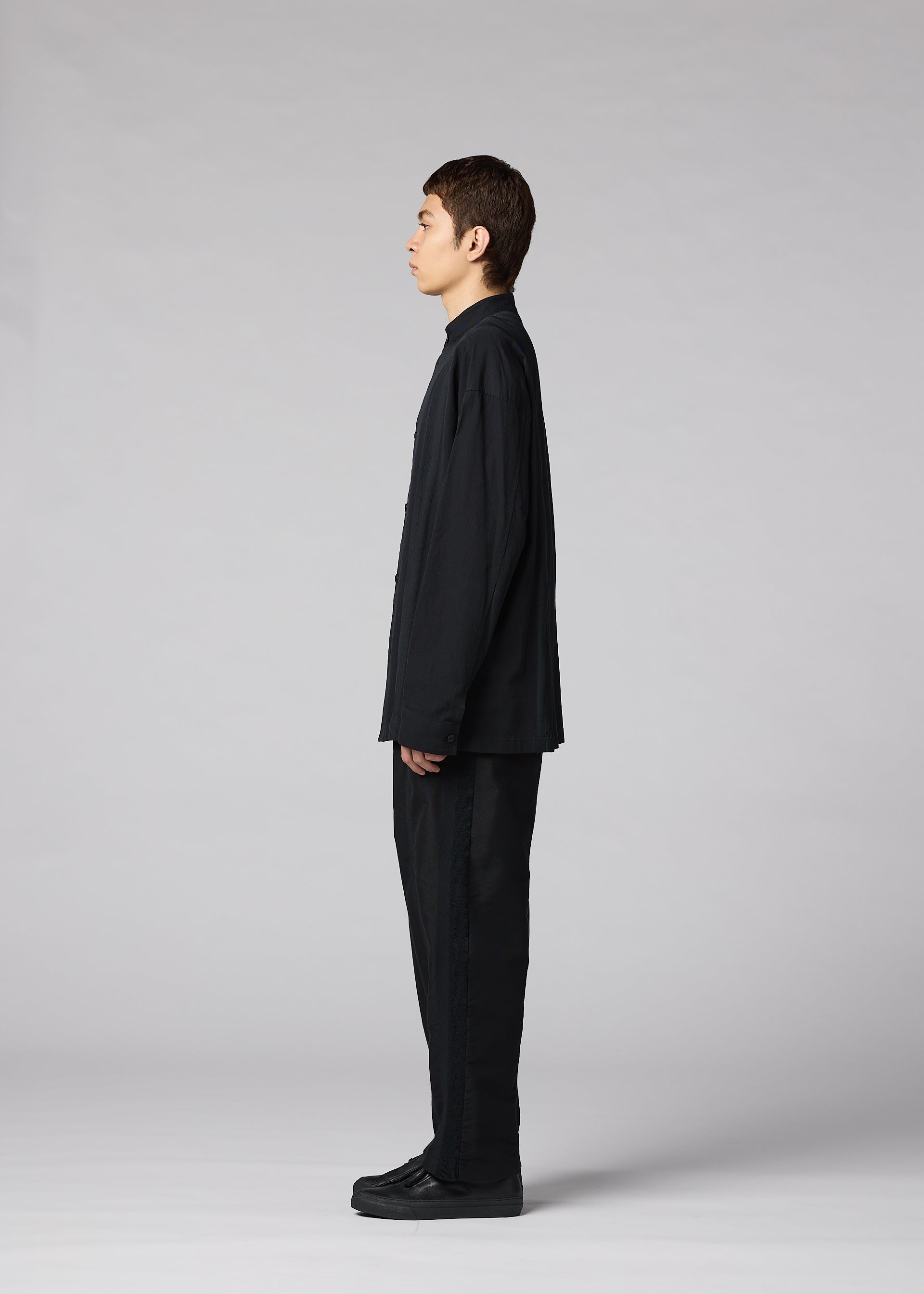 Zoomed view A person stands in profile facing left, wearing the IM MEN CLAY SHIRT—a loose-fitting black cotton stand-up collar shirt—and black cotton pants with black shoes, set against a plain gray background.