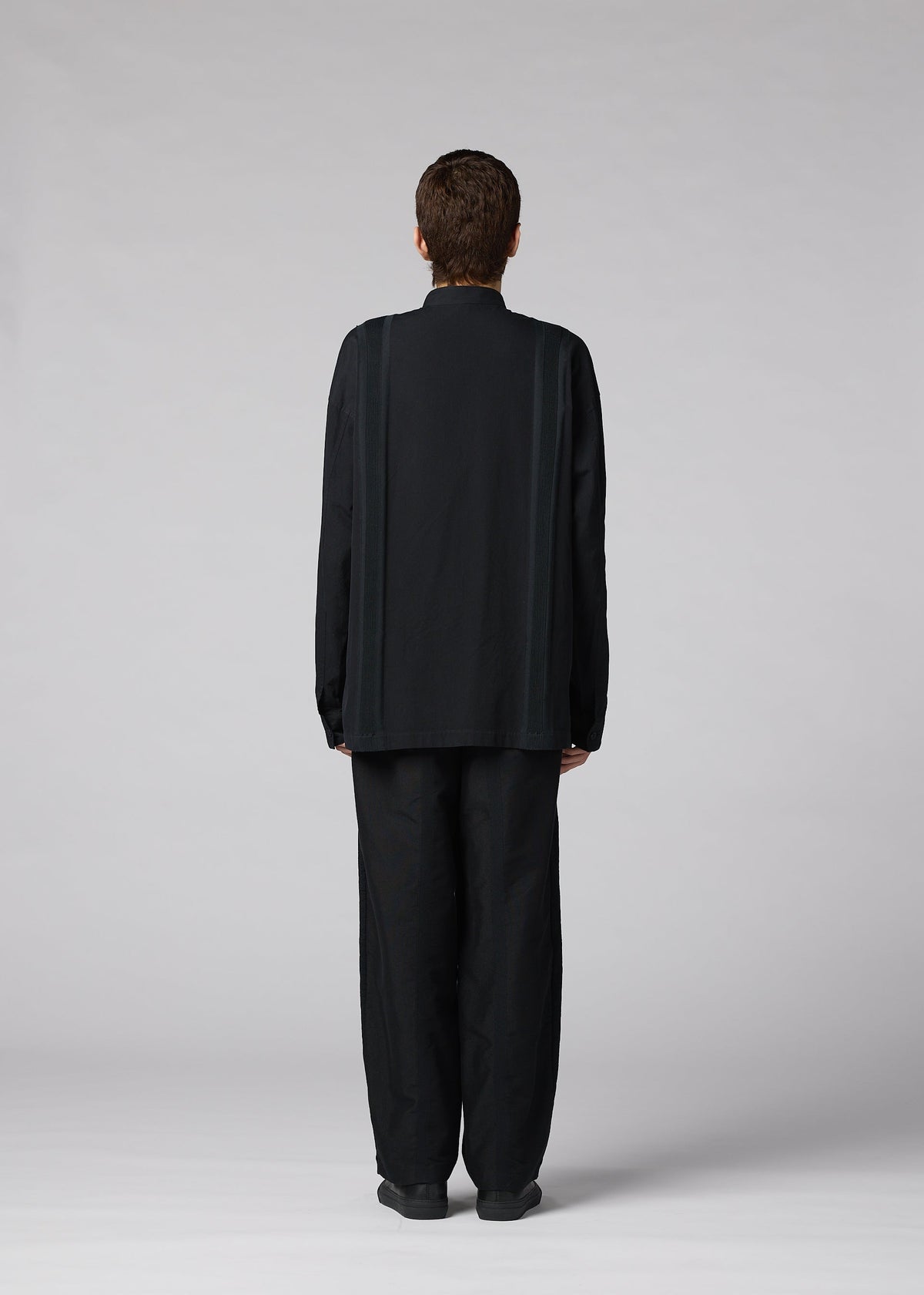 A person with short hair faces away from the camera, wearing the IM MEN CLAY SHIRT— a loose-fitting, long-sleeve stand-up collar shirt— and wide black cotton pants, set against a plain light gray background. view 7 of 8
