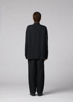 A person with short hair faces away from the camera, wearing the IM MEN CLAY SHIRT— a loose-fitting, long-sleeve stand-up collar shirt— and wide black cotton pants, set against a plain light gray background.