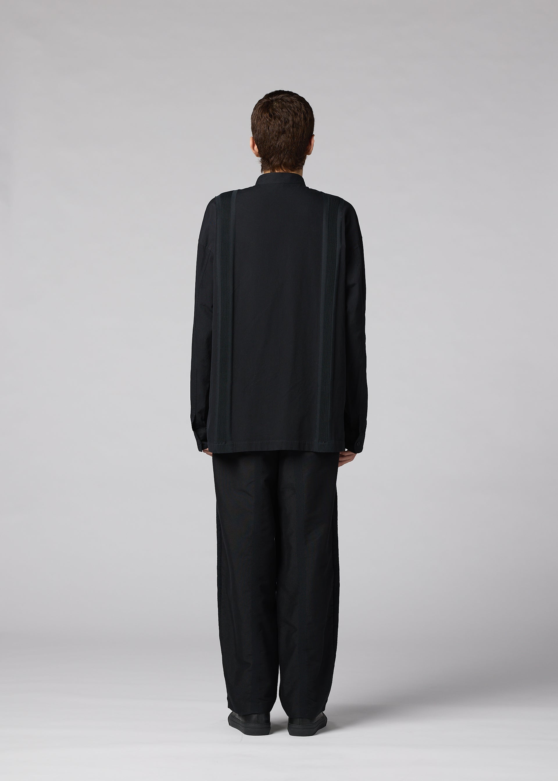 Zoomed view A person with short hair faces away from the camera, wearing the IM MEN CLAY SHIRT— a loose-fitting, long-sleeve stand-up collar shirt— and wide black cotton pants, set against a plain light gray background.
