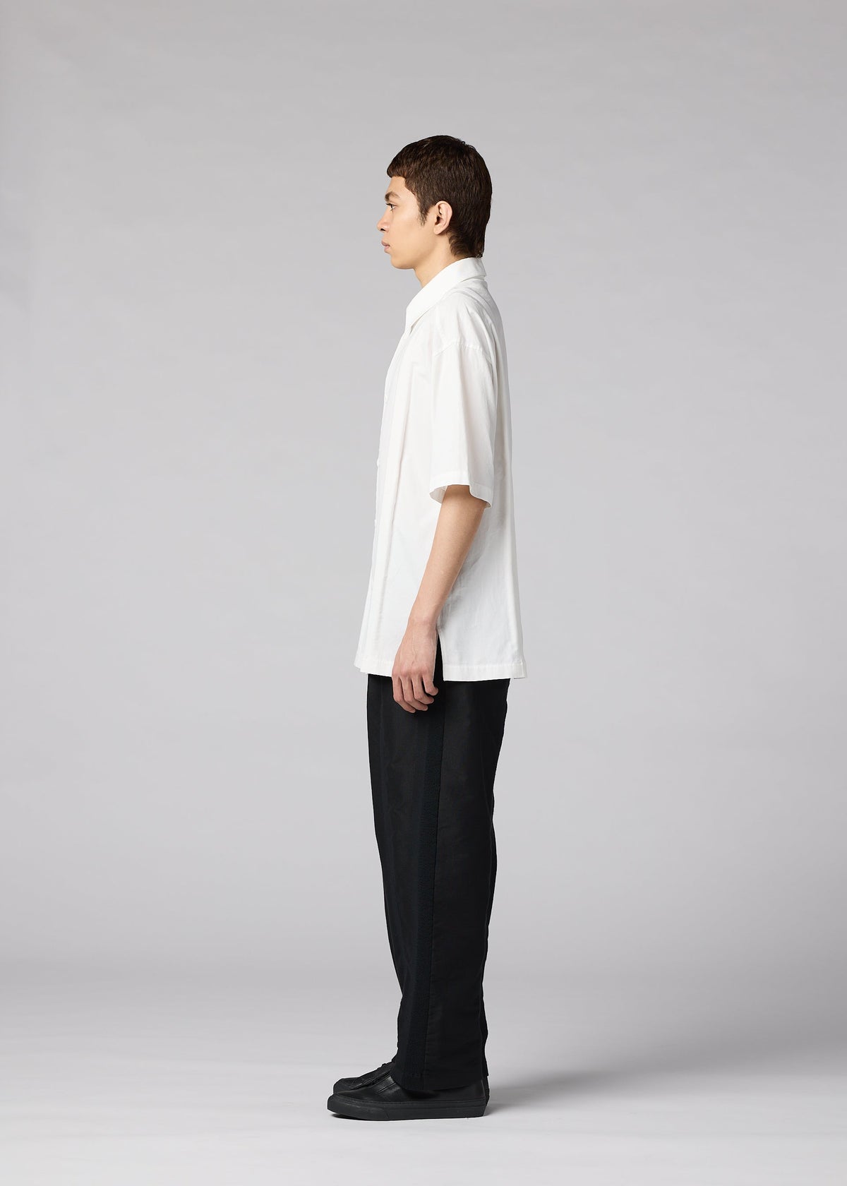 A person with short dark hair stands in profile, facing left, wearing the IM MEN CLAY SHIRT (loose white cotton, short sleeves), black pants, and black shoes against a plain light gray background. view 6 of 8